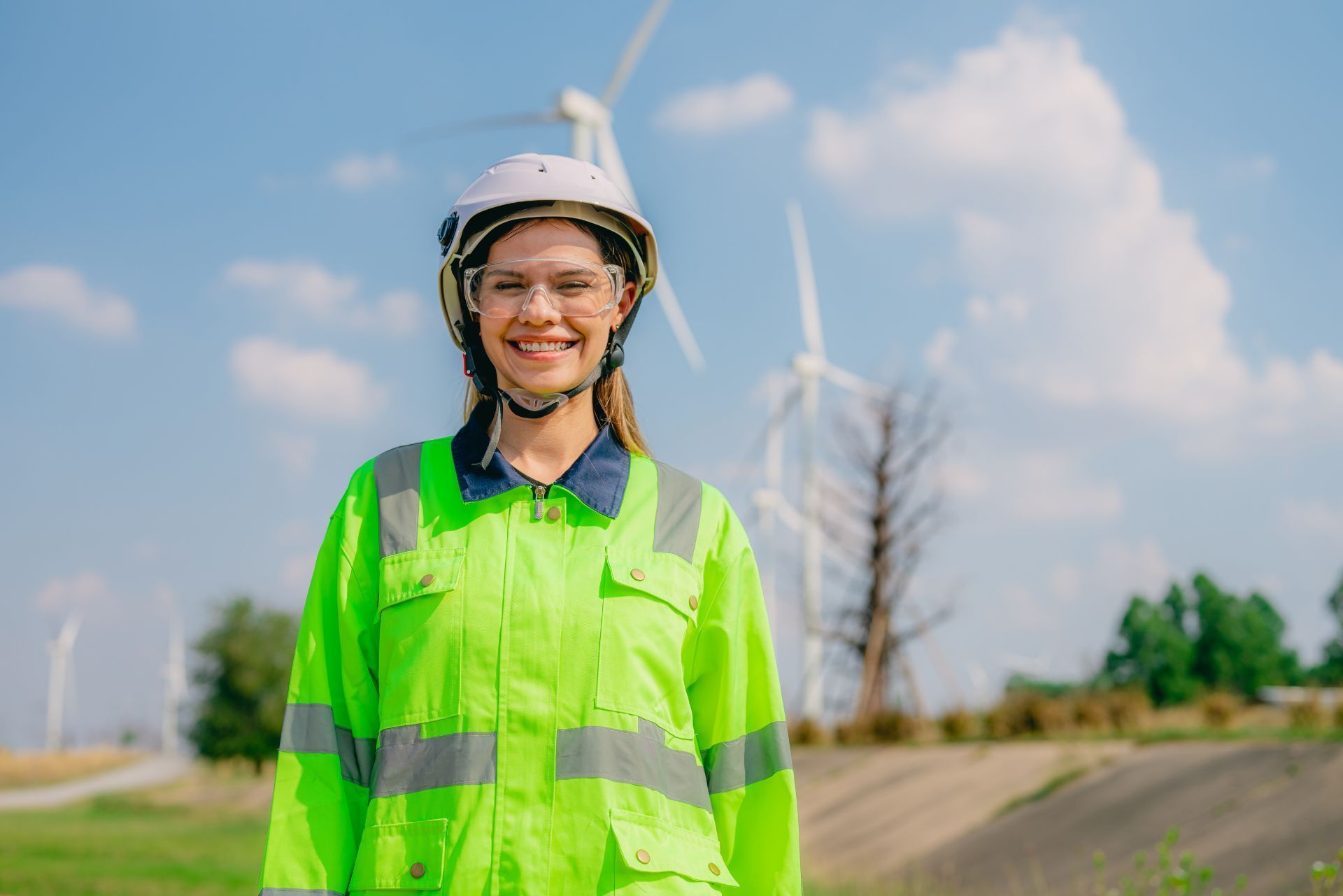 A smiling worker in a neon high-visibility jacket, safety glasses, and a hard hat standing in a wind turbine field.