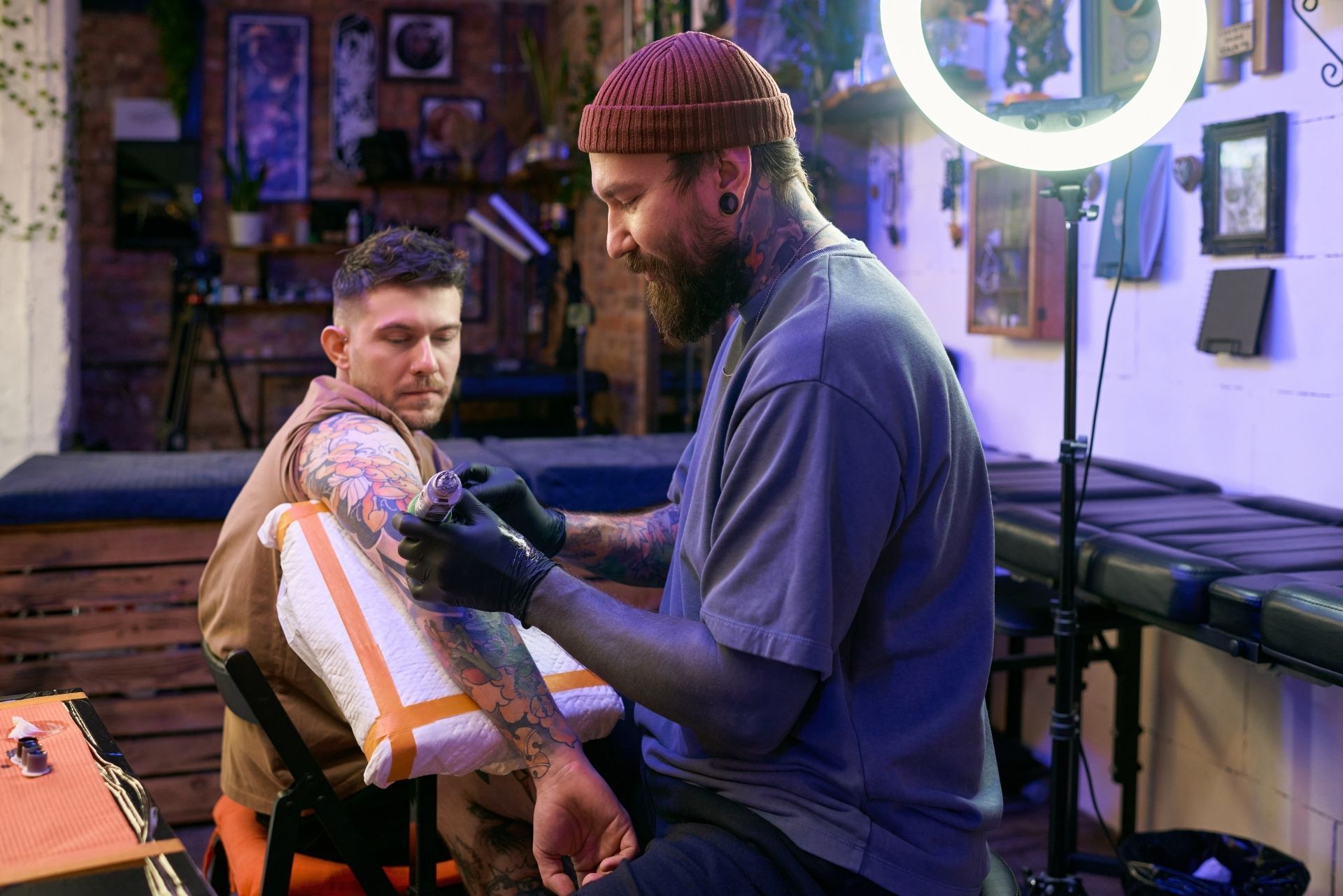 Tattoo artist applying ink to a client’s arm in a studio with a neon ring light and tattoo chair.