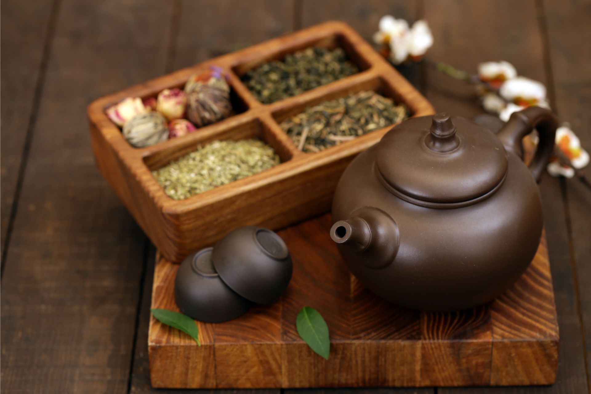 Brown teapot and tea cups on a wooden tray beside a box of loose tea leaves and flowers