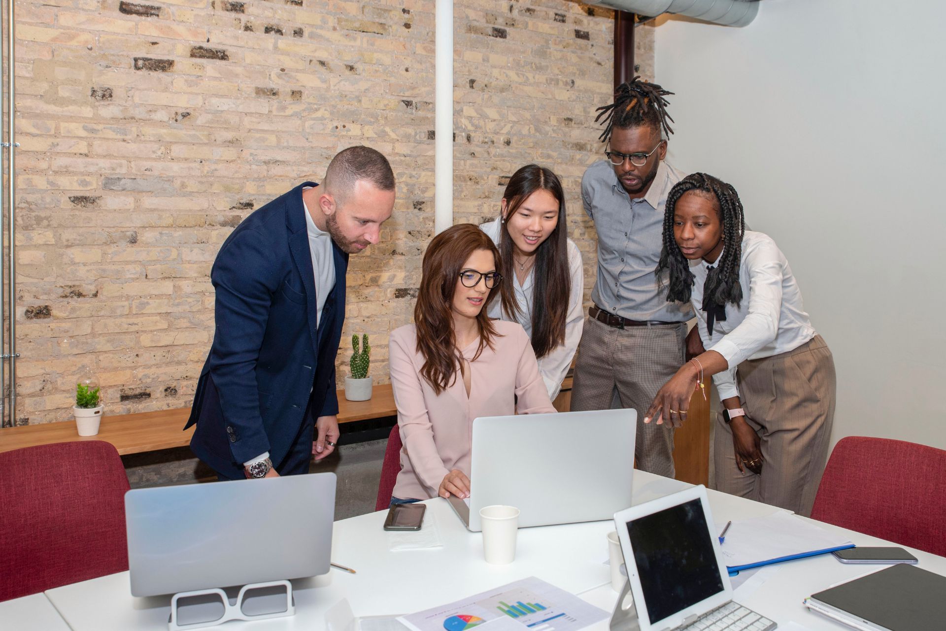A diverse group of colleagues collaborate around a laptop in an office with a brick wall.