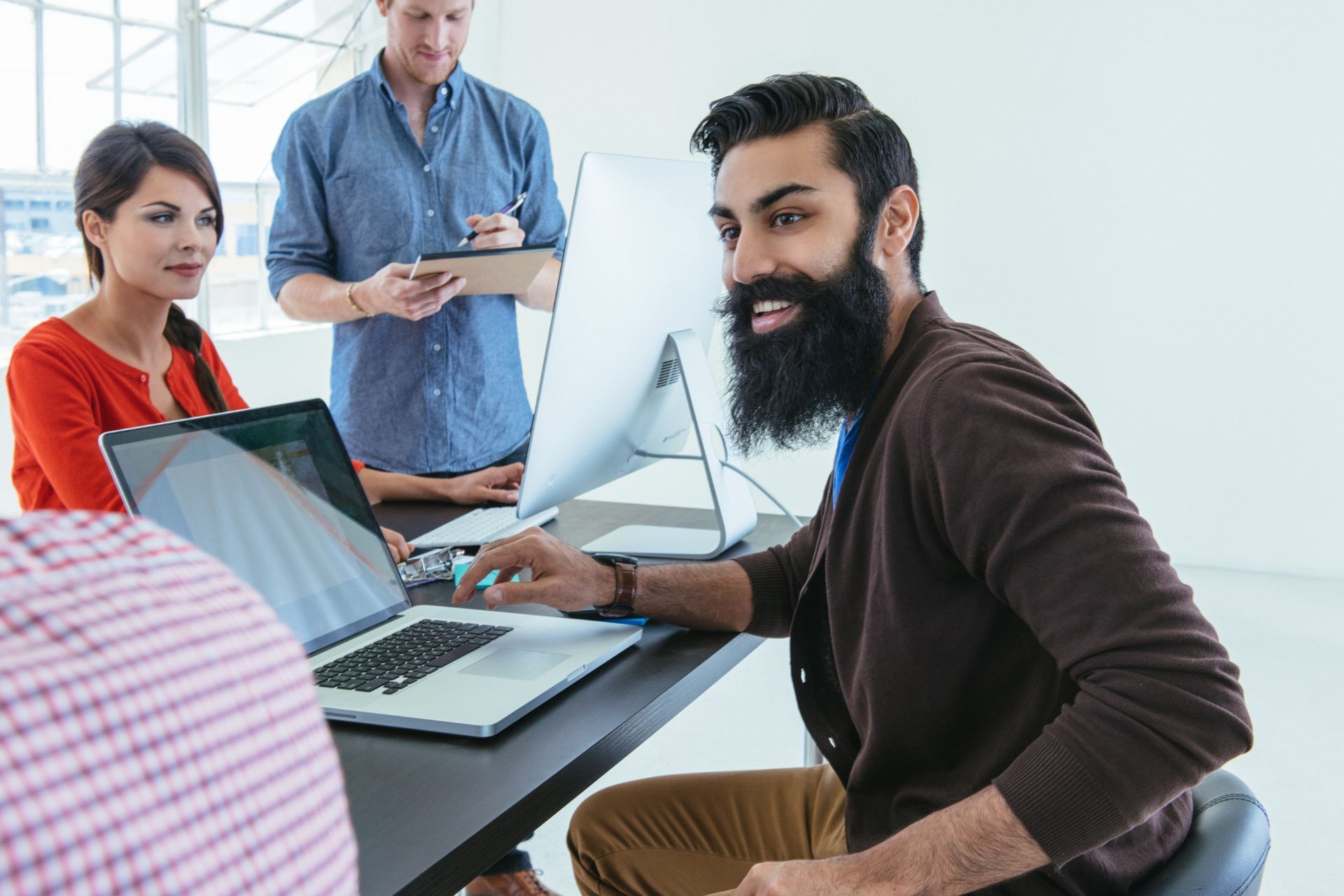 A group of colleagues collaborate in a modern office, focusing on laptops and a desktop computer.