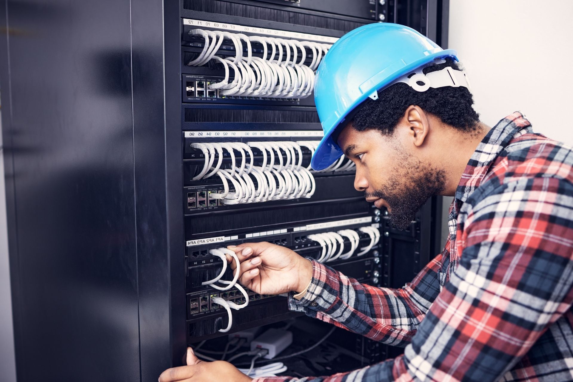 A technician in a blue hard hat and plaid shirt inspects white cables in a black server rack.