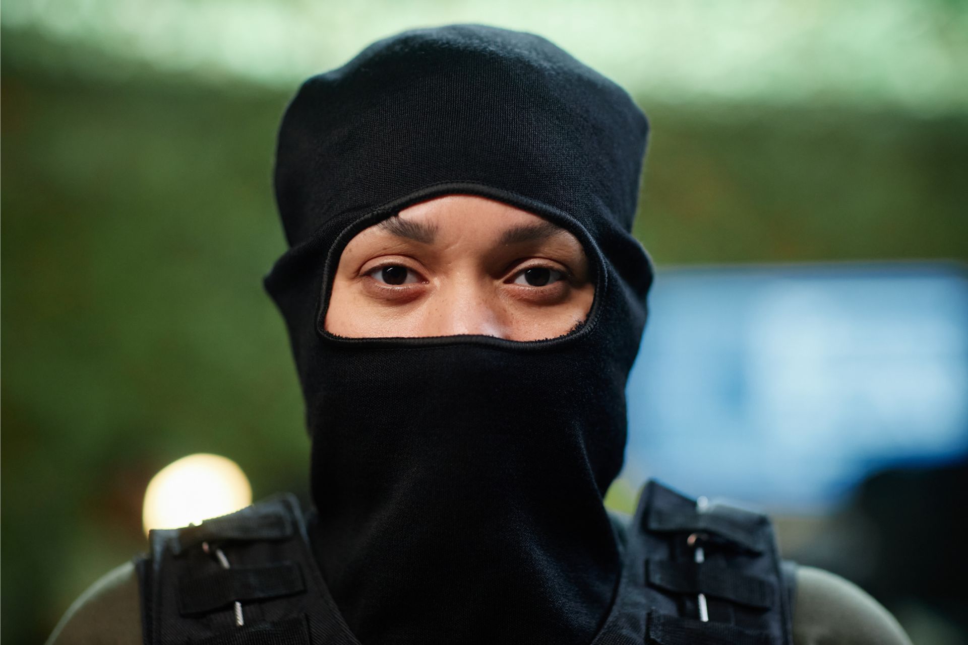 Person in black tactical clothing and balaclava, looking at the camera against a blurred outdoor background.