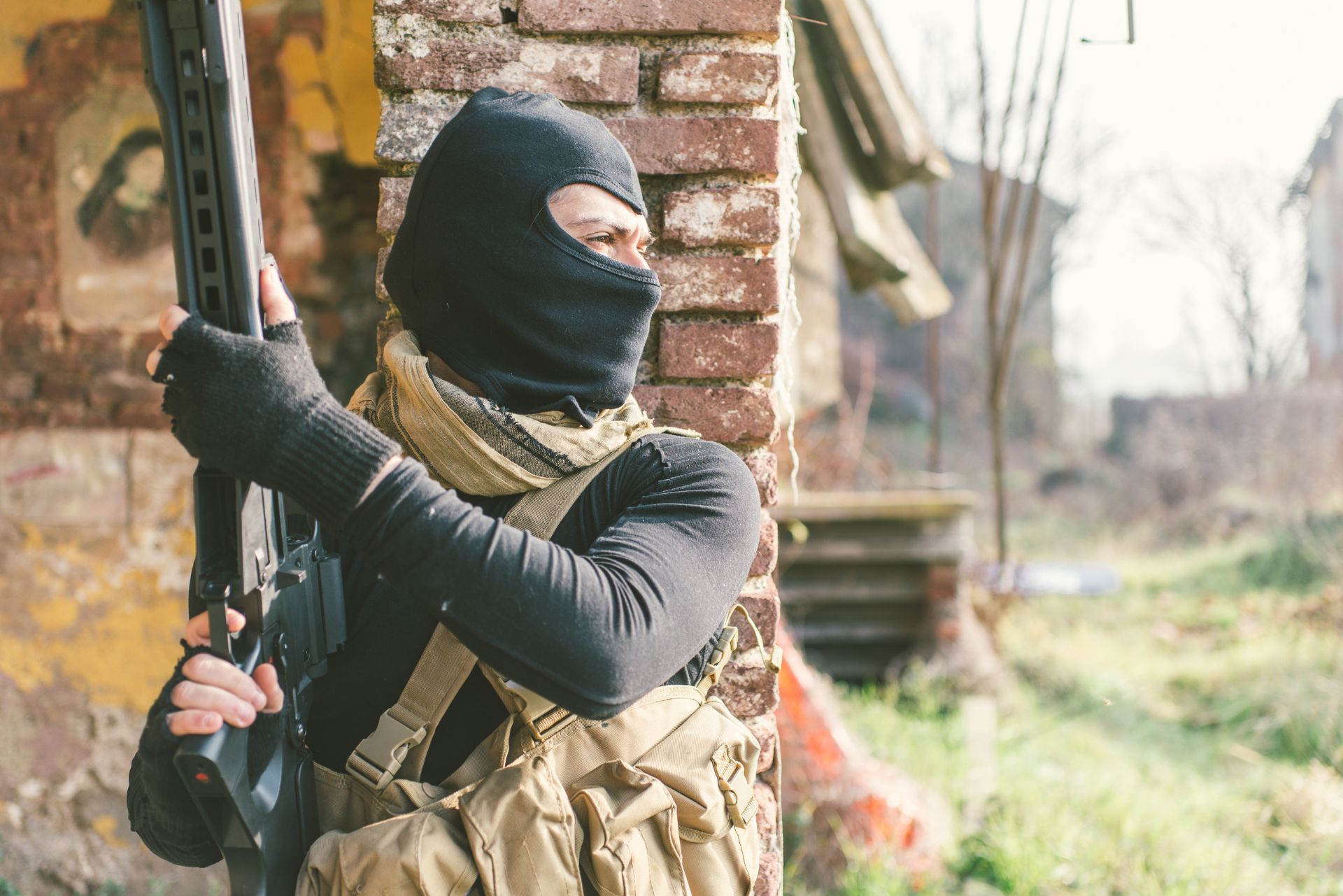 Masked person holding a rifle beside a brick wall, scanning outdoors in camouflage gear