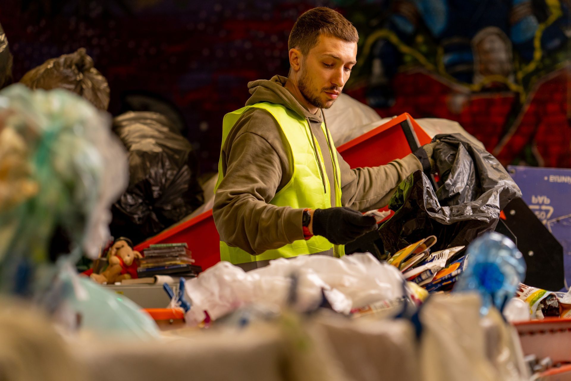 A worker in a high-visibility vest sorts through various discarded items and recyclables on a conveyor belt.