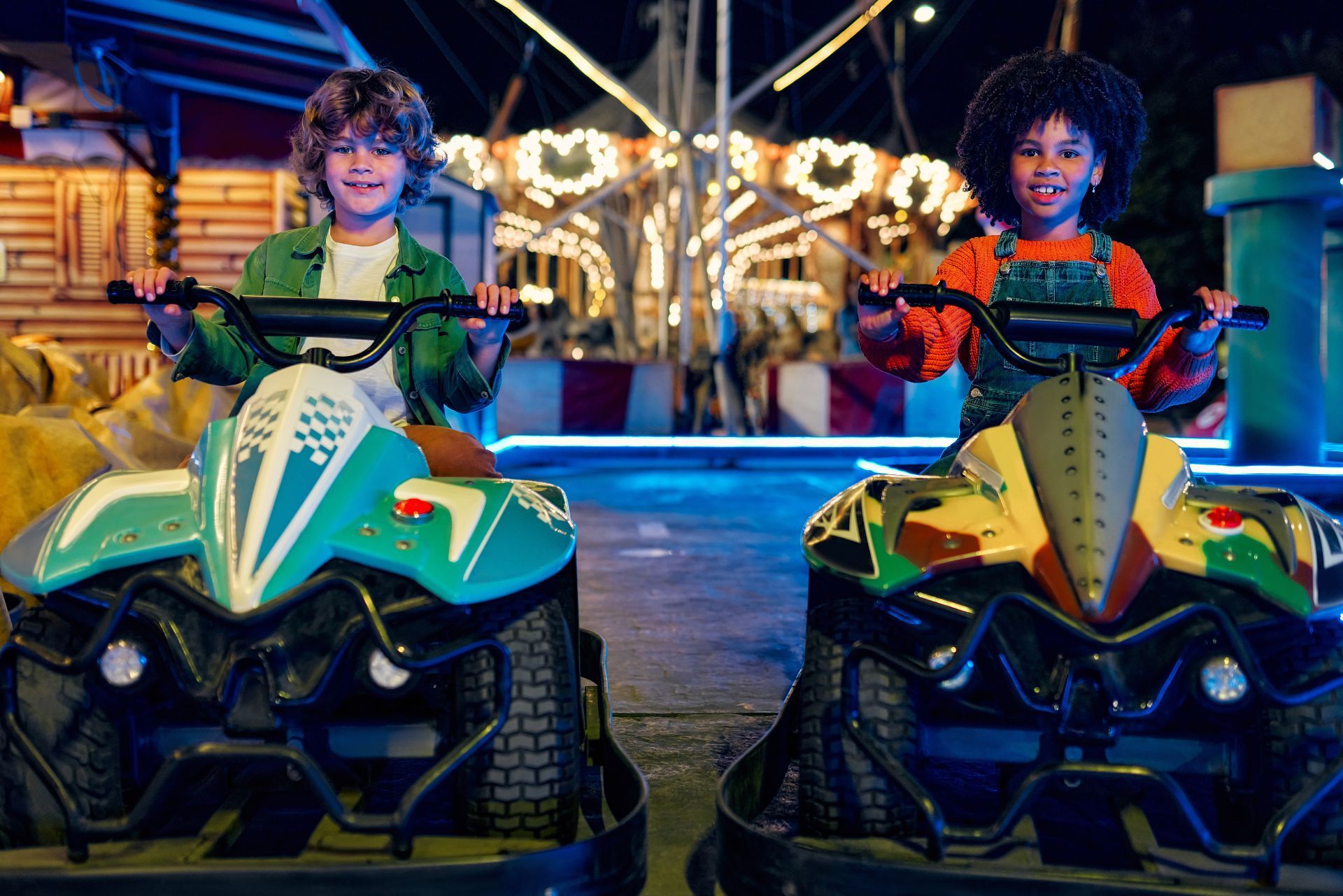 Two children ride colorful ATVs at a fairground with a lit-up carousel in the background.