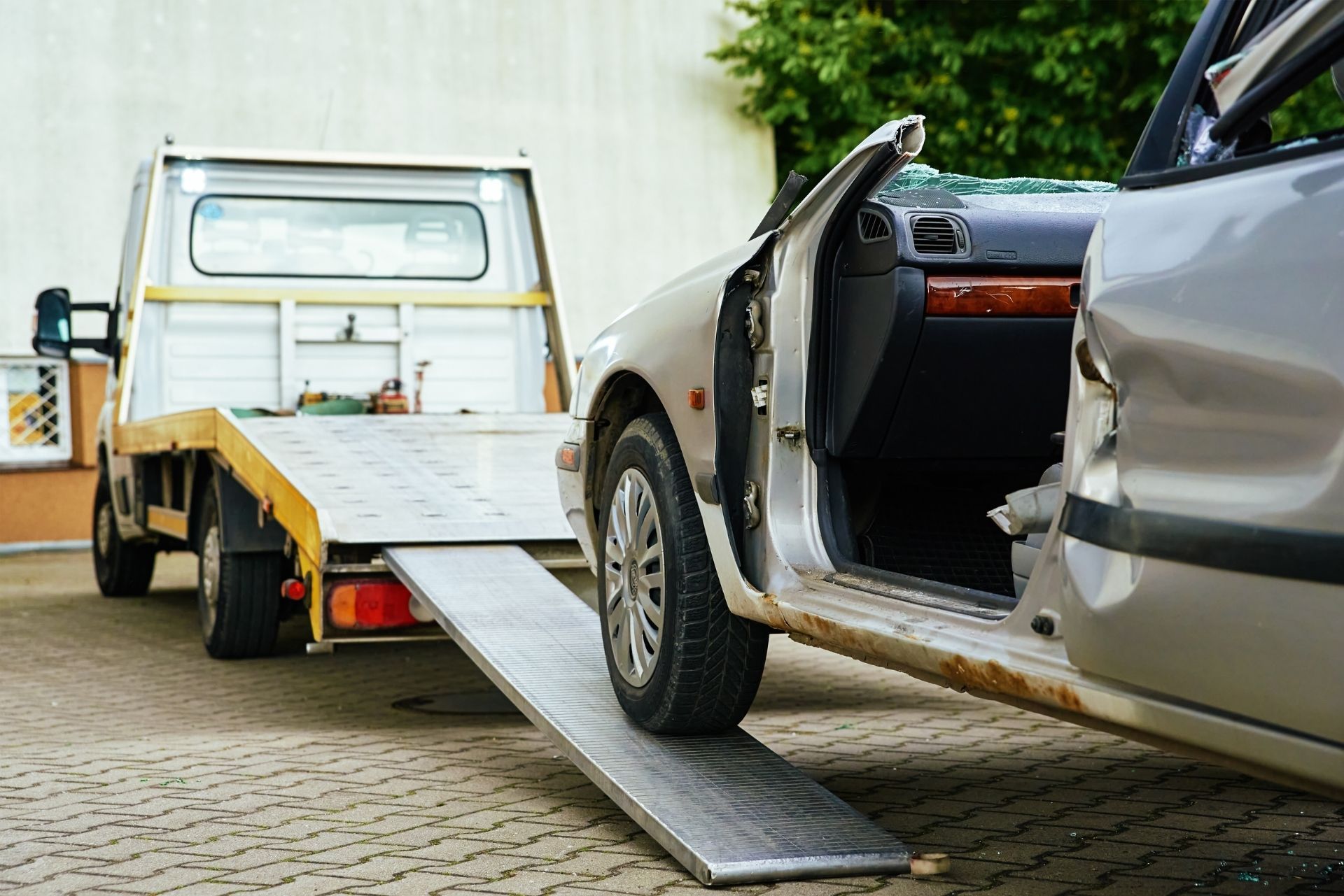 Silver car being loaded onto a yellow tow truck with a ramp on a cobblestone street