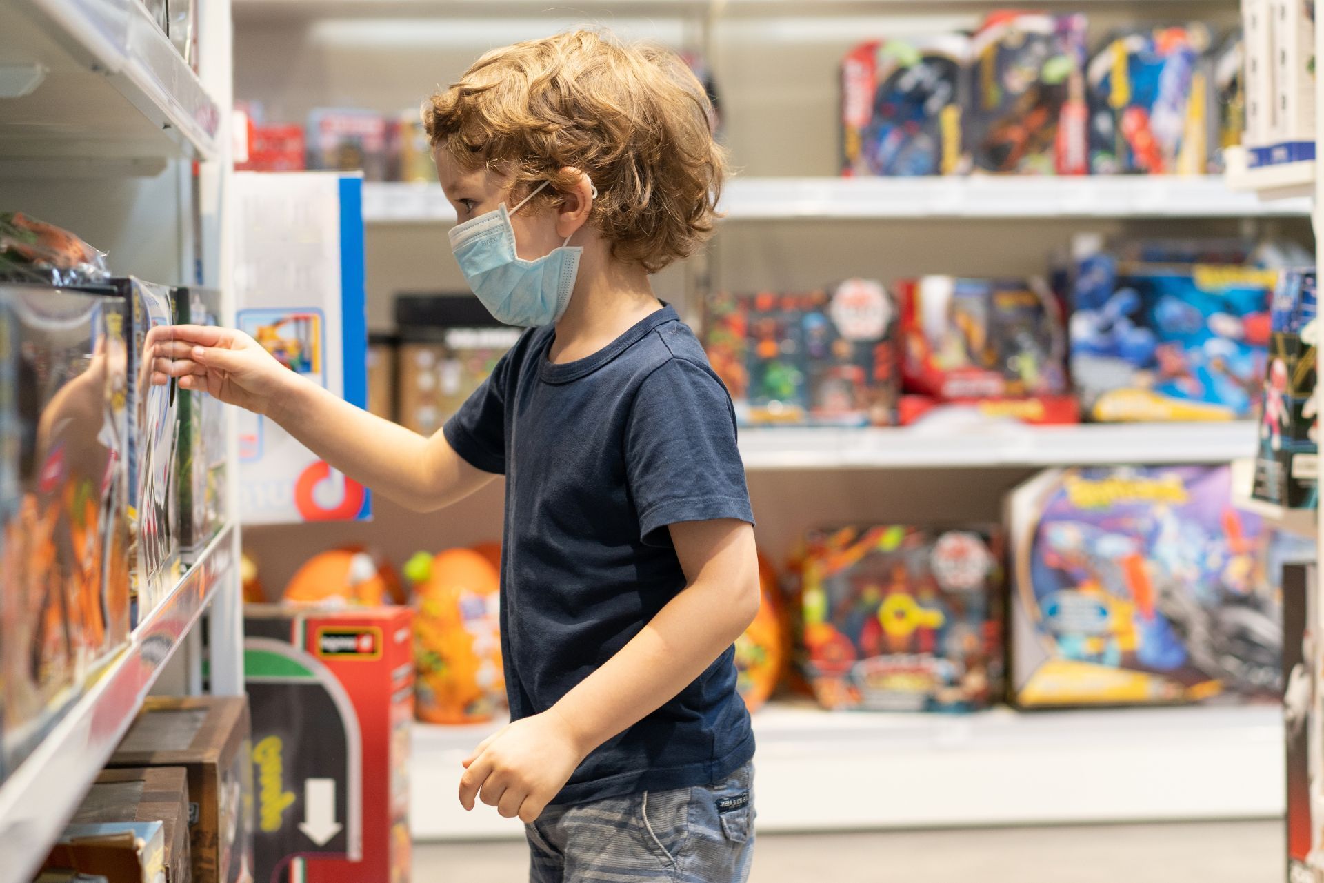 Child in a store aisle selecting a toy from a shelf, wearing a blue mask and navy shirt.
