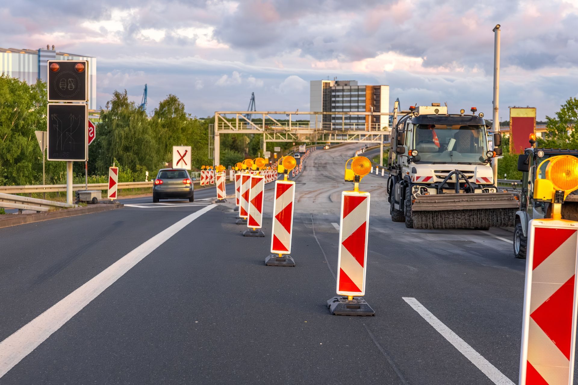 A road construction site with red and white barricades lining the asphalt, near a truck and a moving car at sunset.