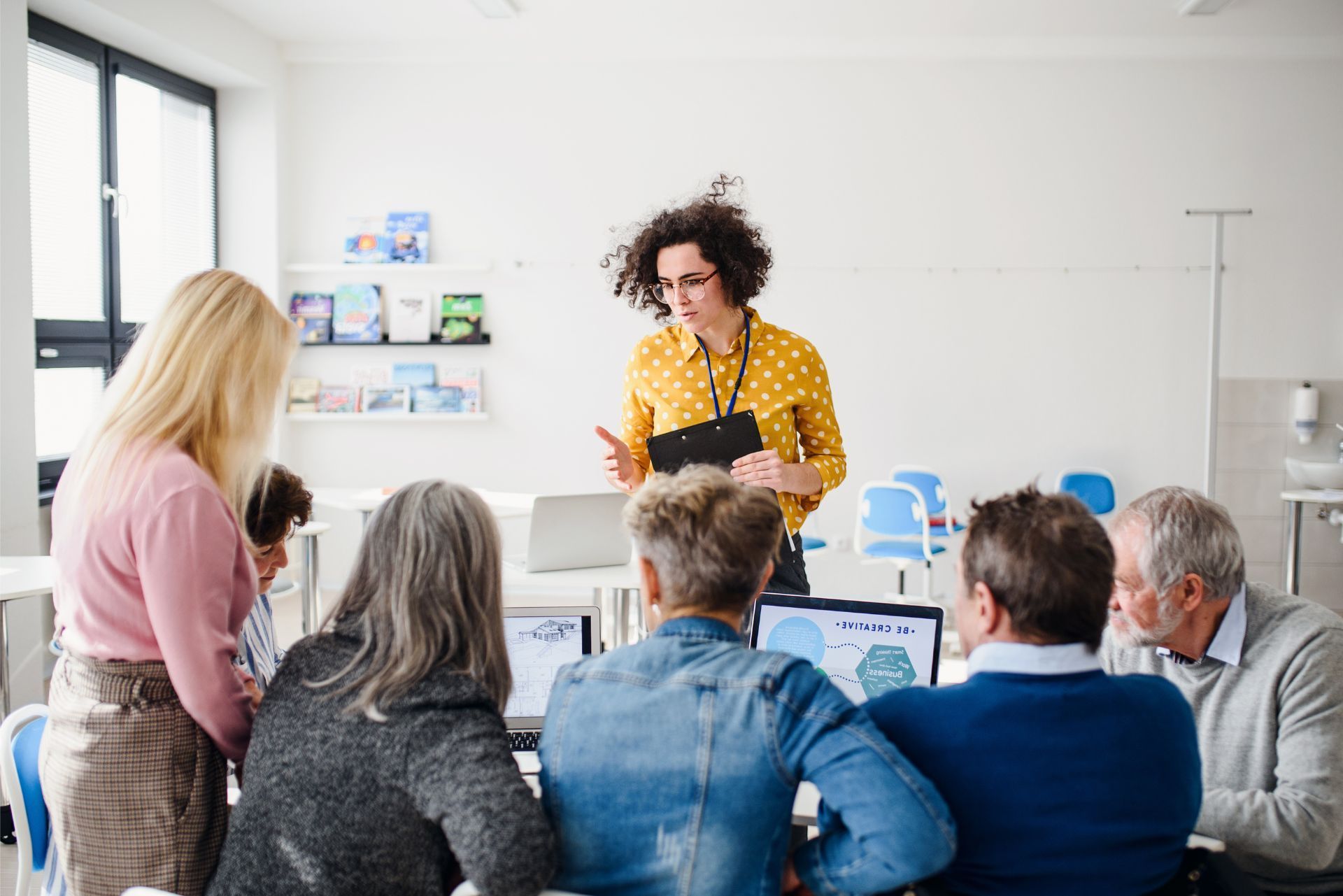 A woman in a yellow shirt leads a workshop for a group of people sitting around tables with laptops in a bright room.