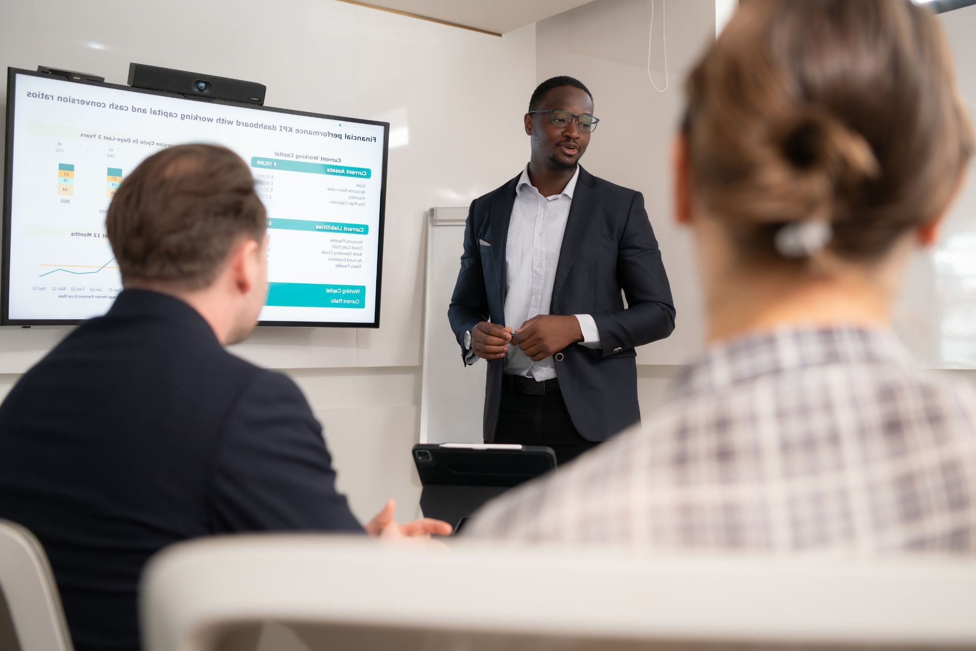A person in a suit presents a digital data chart to two seated colleagues in an office setting.