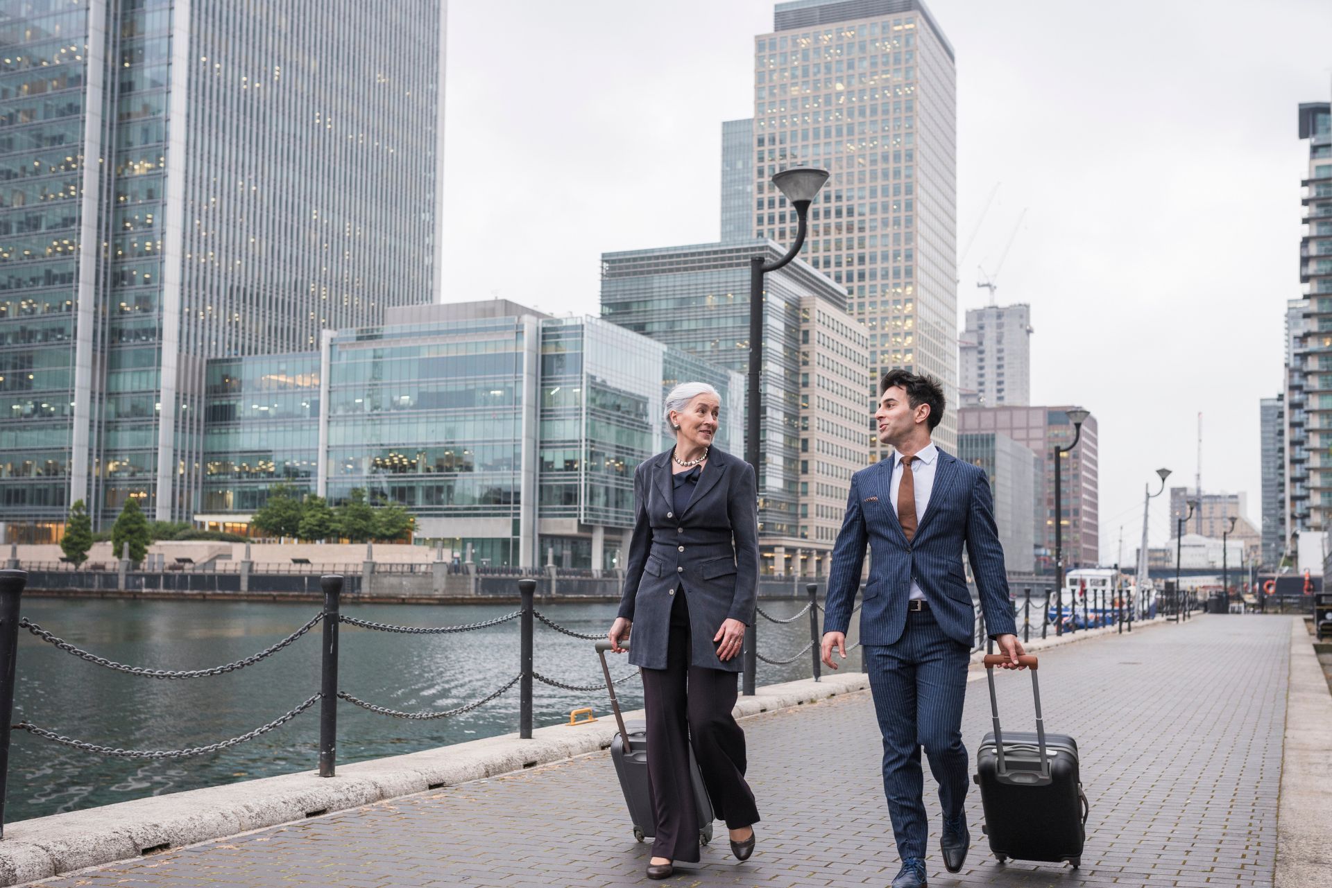 Two business travelers walking beside a waterfront city skyline, one pulling a rolling suitcase.