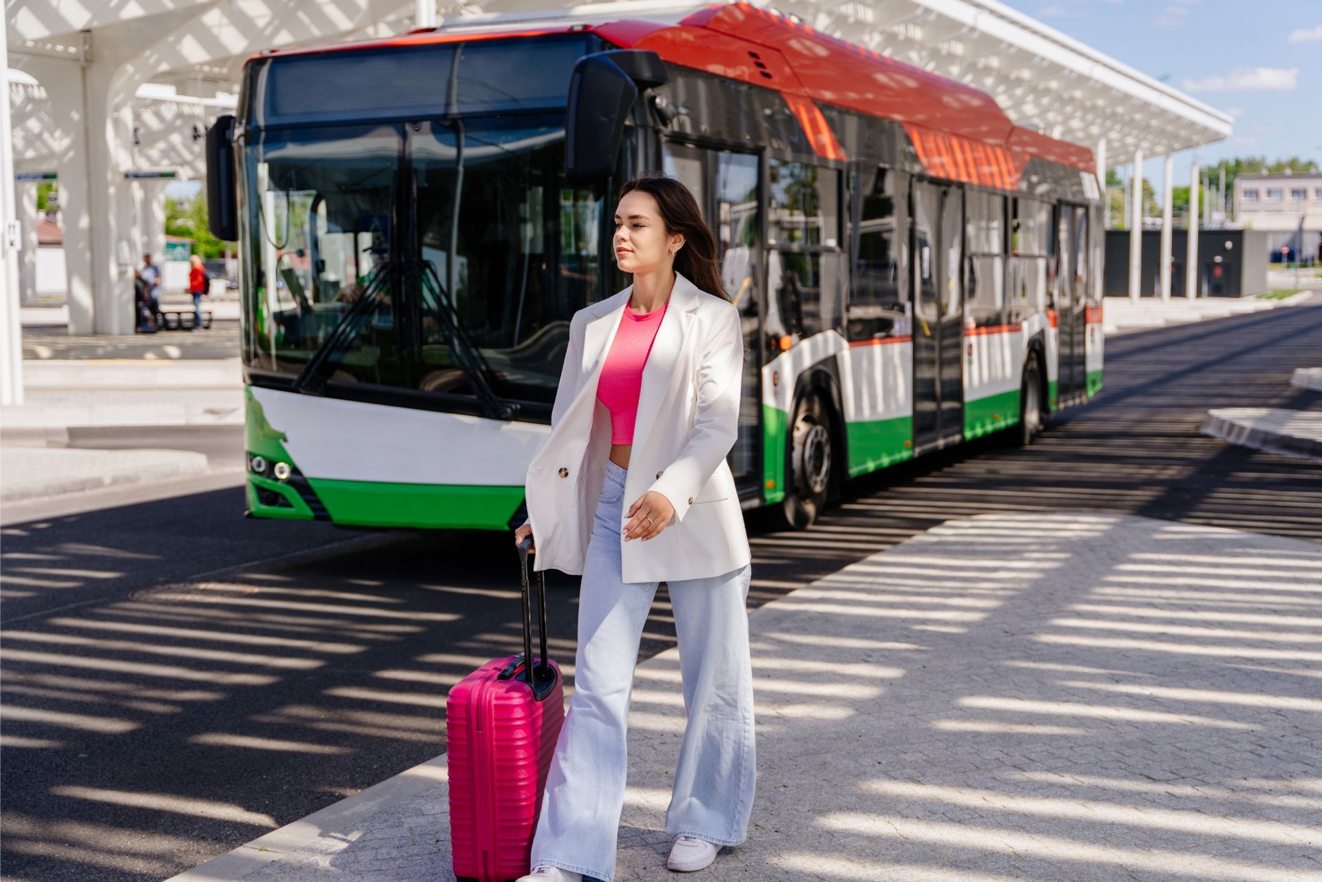 A traveler with a pink suitcase walks near a parked bus under a modern, white-pillared terminal structure.