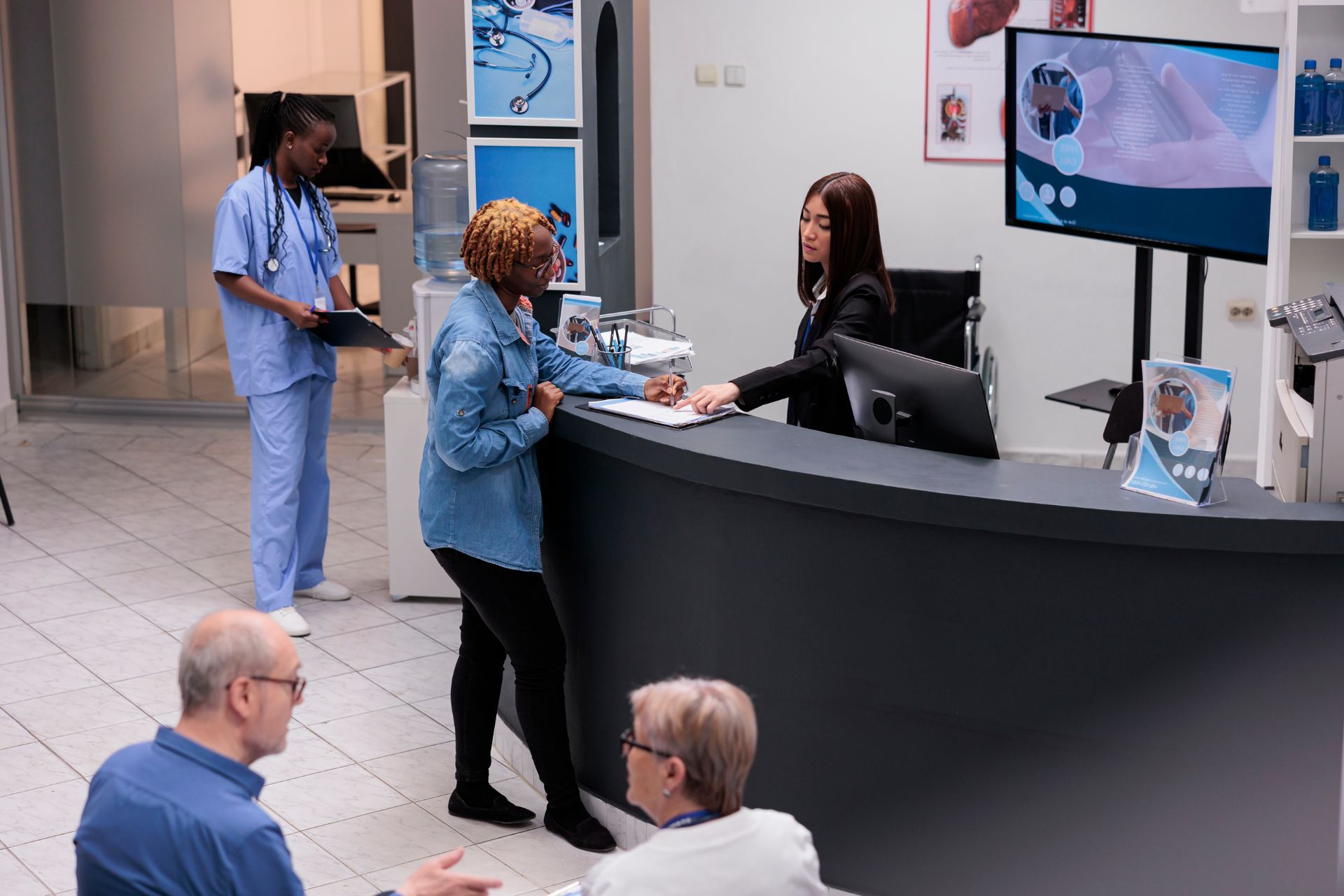 A reception area in a medical facility with staff assisting a patient at a counter while others wait in the foreground.