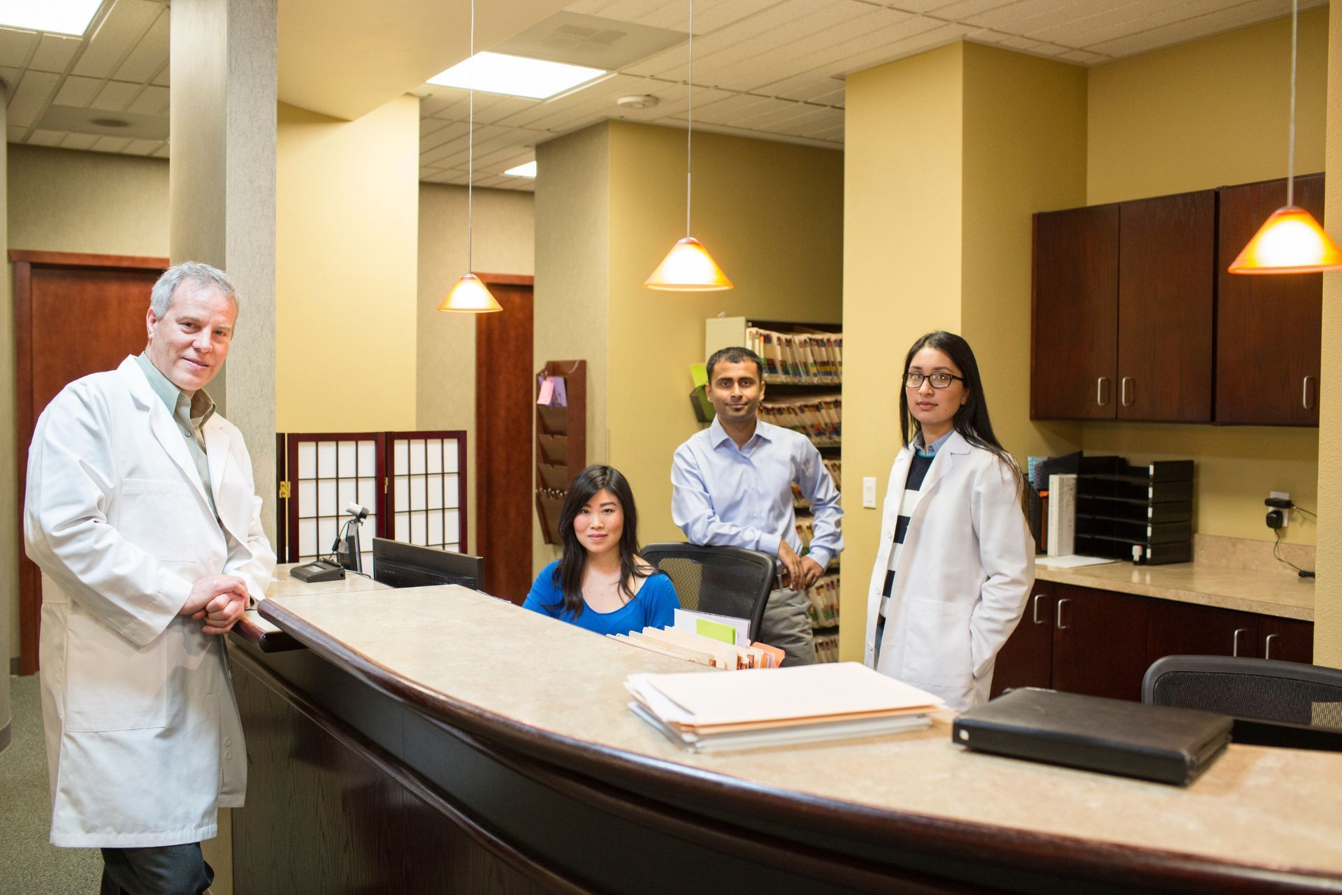 Four medical staff stand behind a reception desk in a professional office setting with cabinets and pendant lighting.