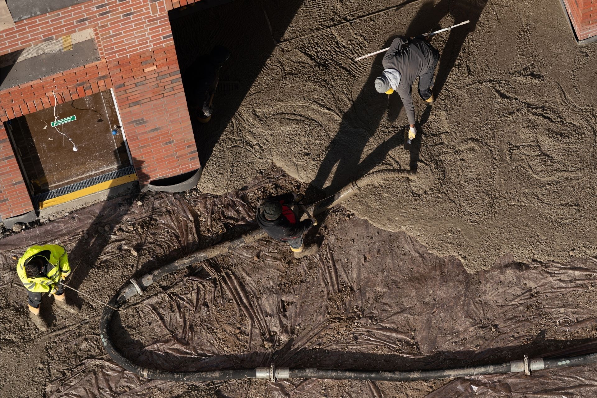 Workers near a brick building guide a hose across a dirt construction site.