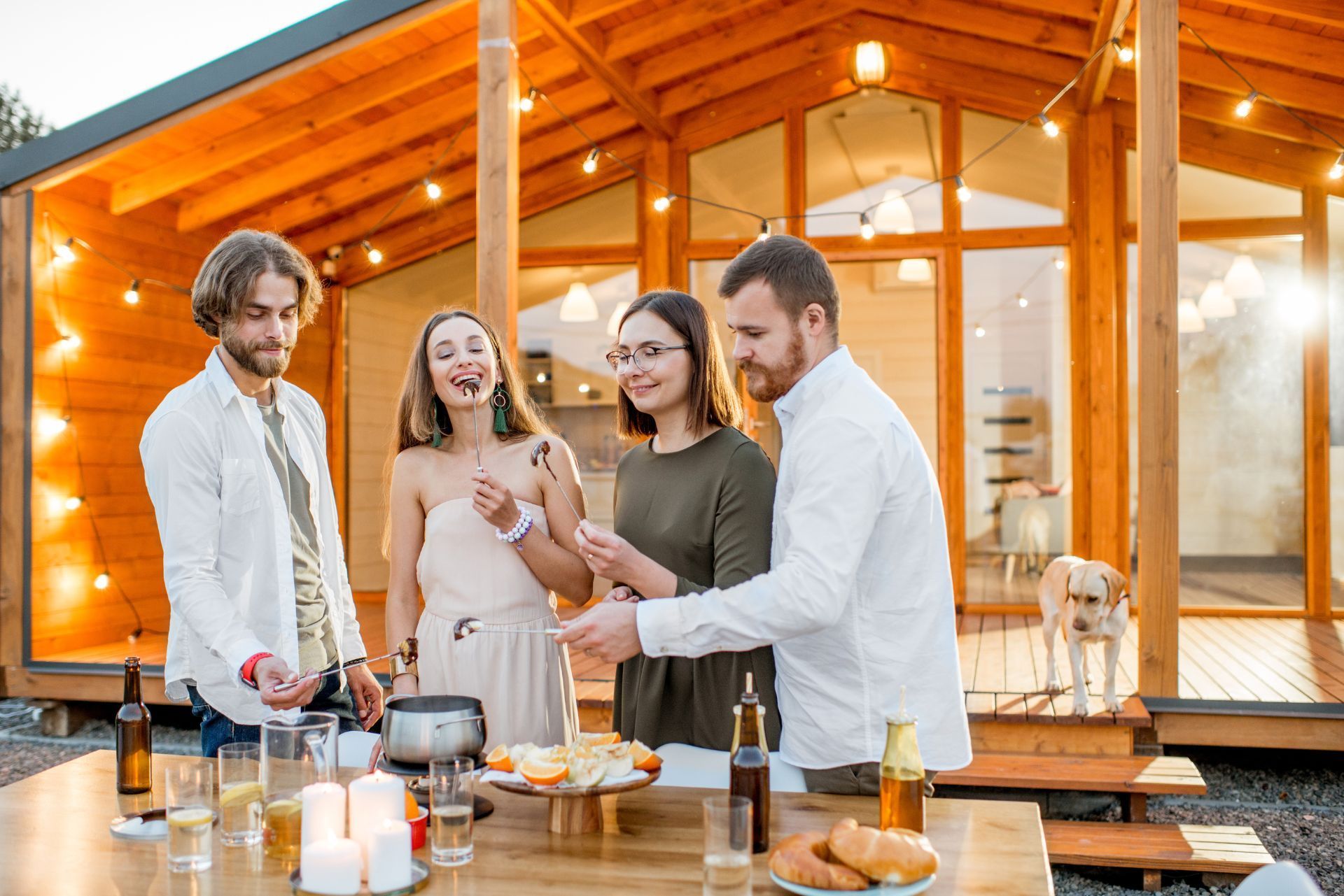 Friends toasting at an outdoor dinner table in front of a warmly lit wooden cabin