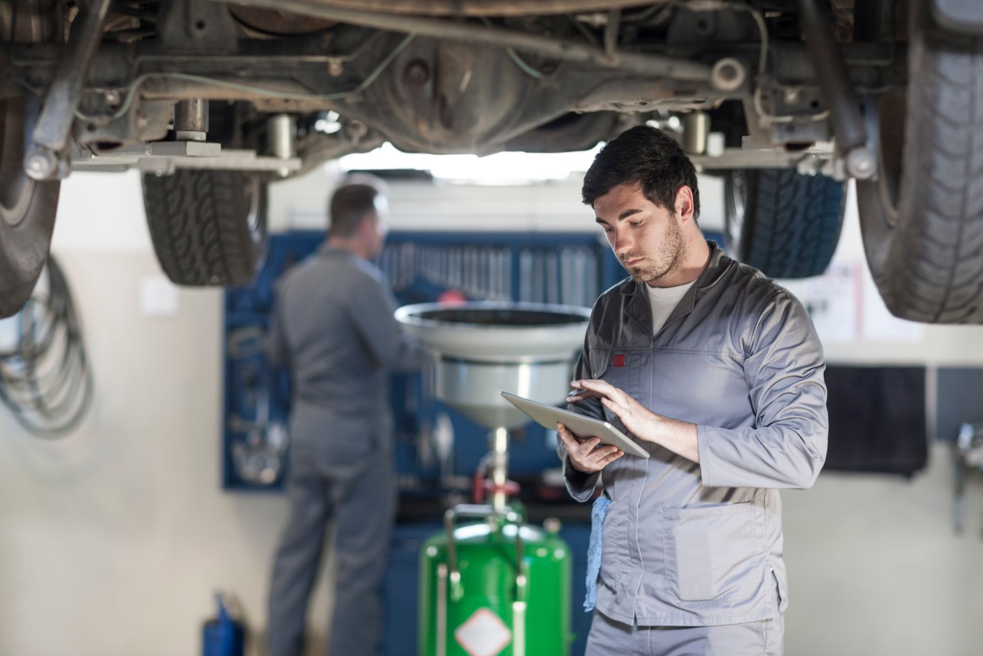 Mechanic in gray coveralls reading a clipboard under a car in an auto repair shop