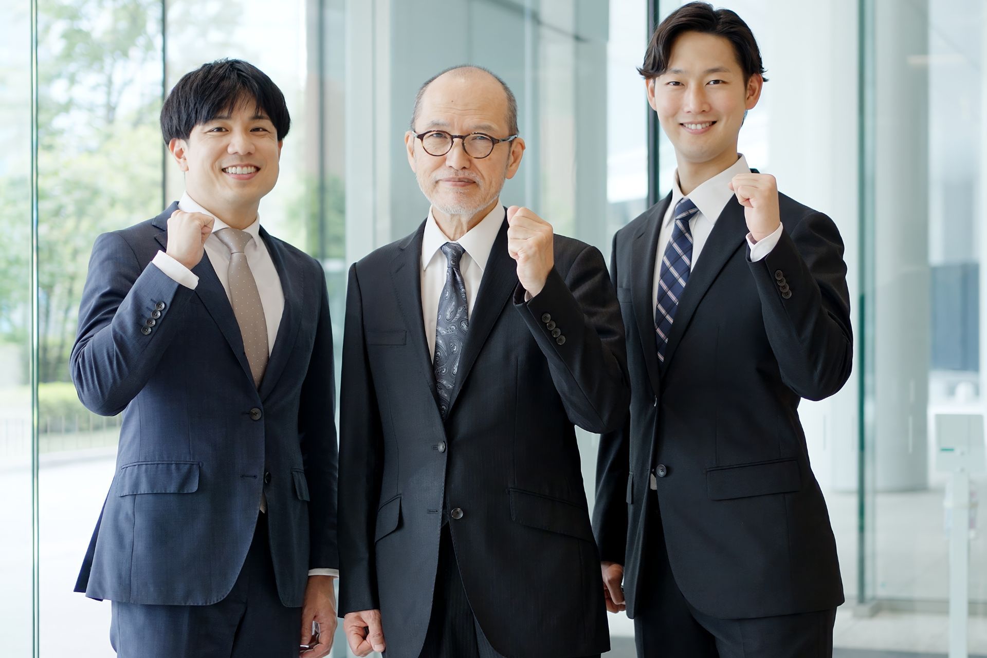 Three businessmen in suits standing together, smiling and raising fists in a bright office lobby