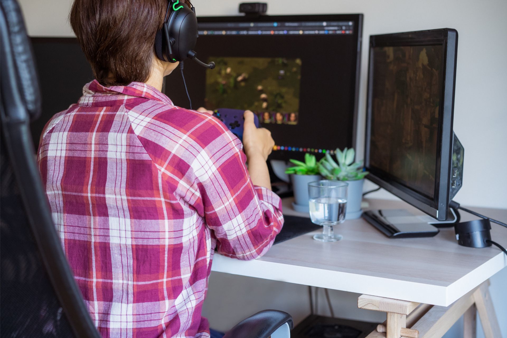 A person in a red plaid shirt wearing a headset plays a video game on a multi-monitor computer setup.