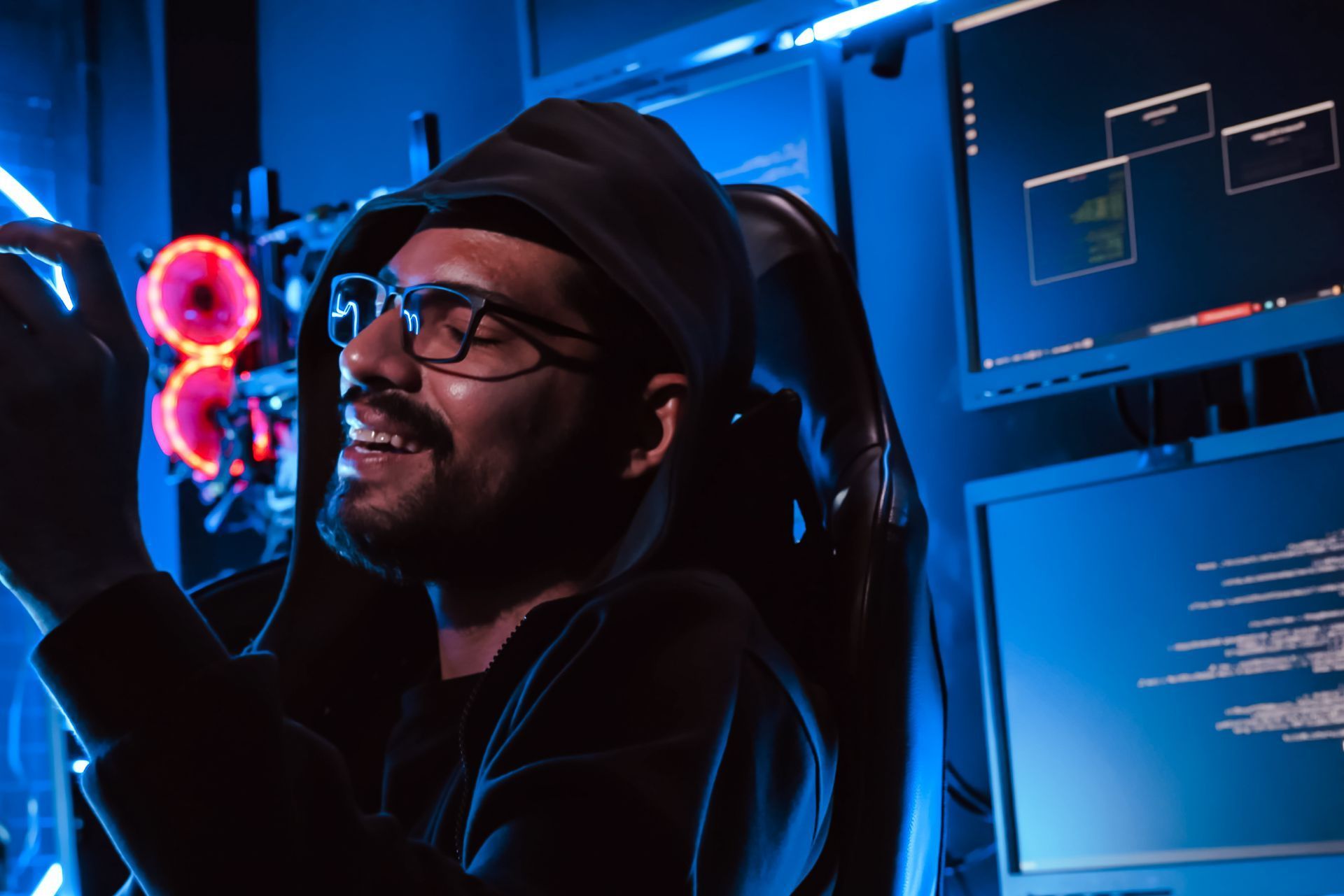 A person in a hooded sweatshirt and glasses works in a dark, blue-lit room with monitors displaying lines of code.