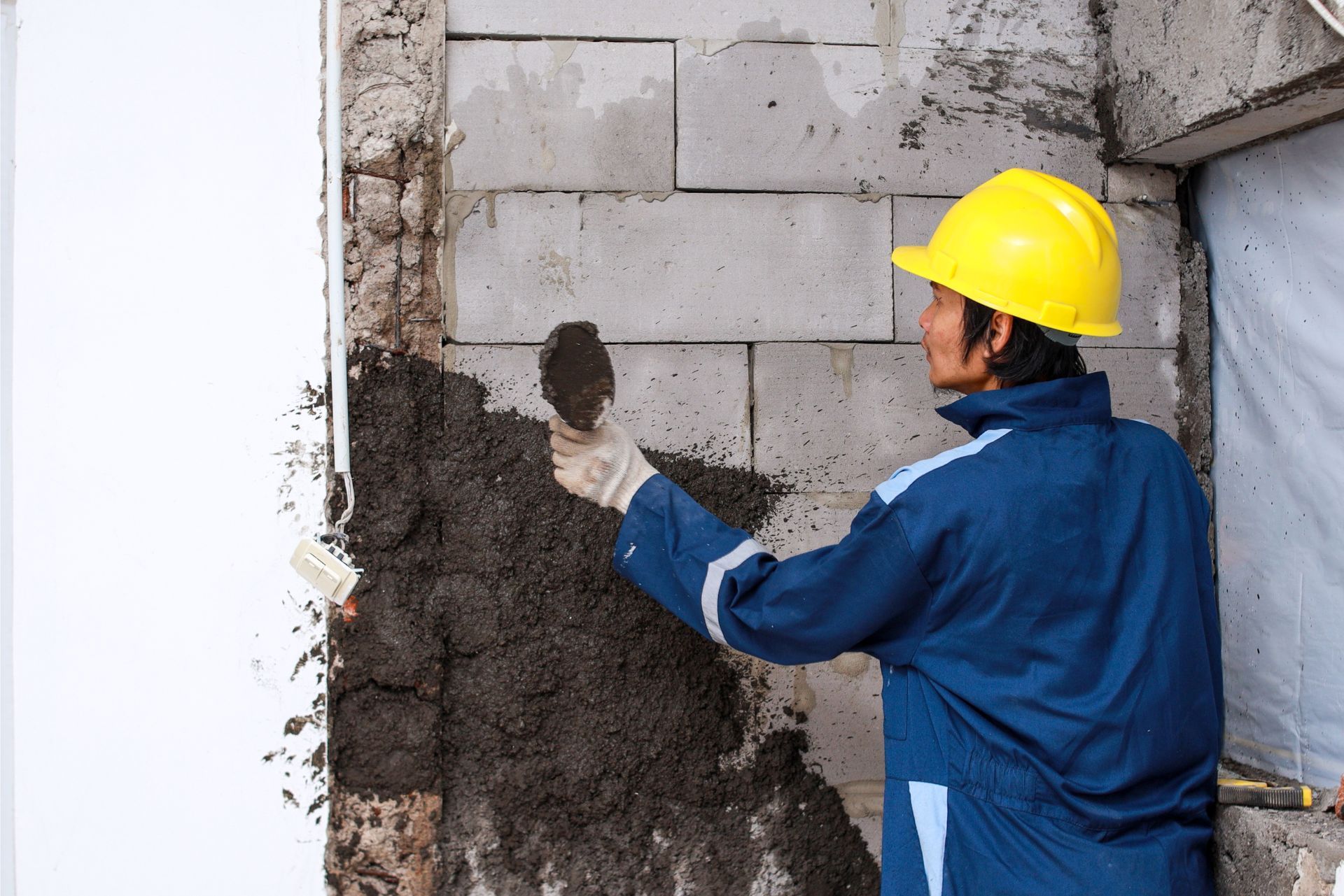 Worker in yellow hard hat applying mortar to a concrete wall seam indoors.