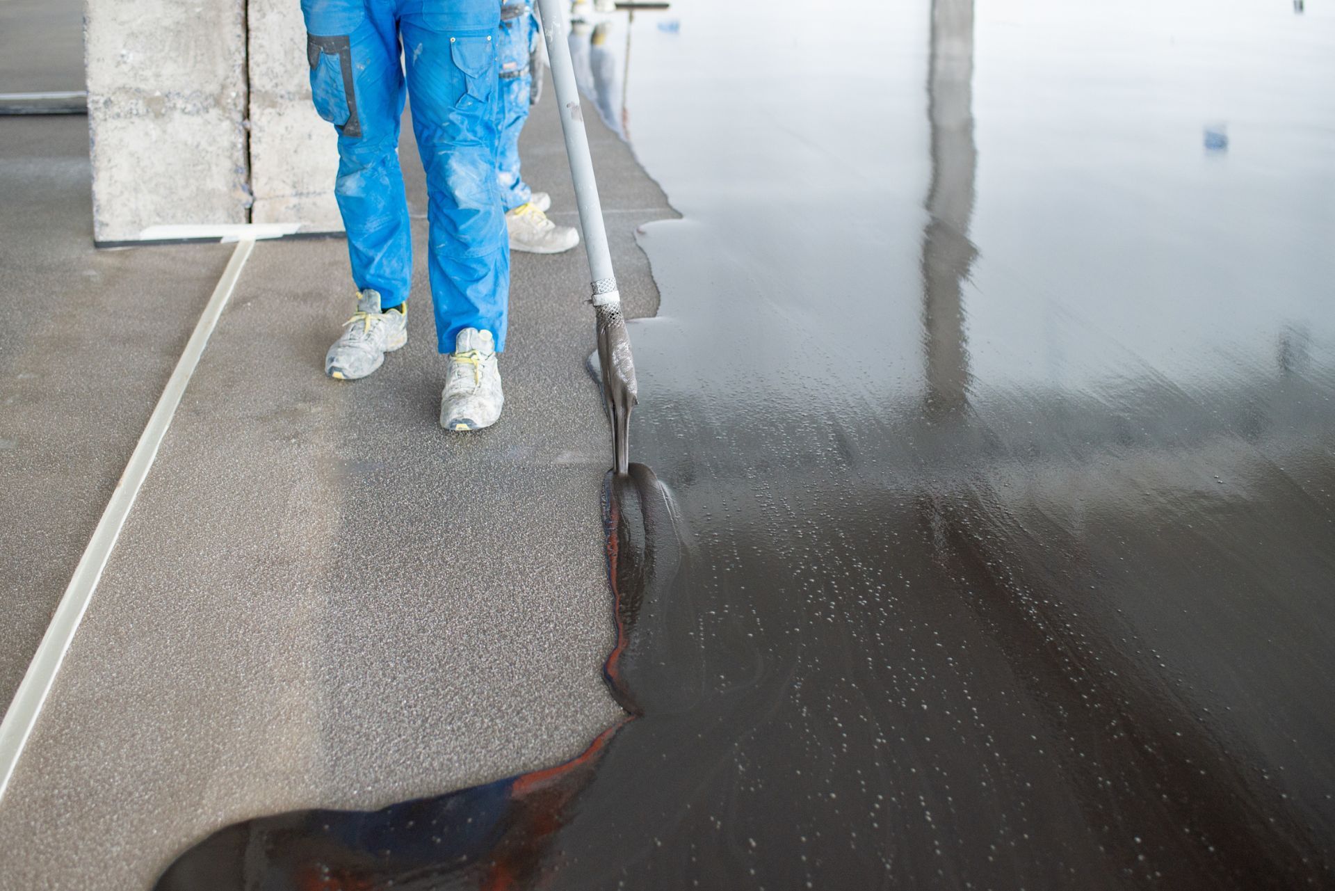 Person in blue coveralls and white boots standing beside wet pavement and a freshly coated dark surface.