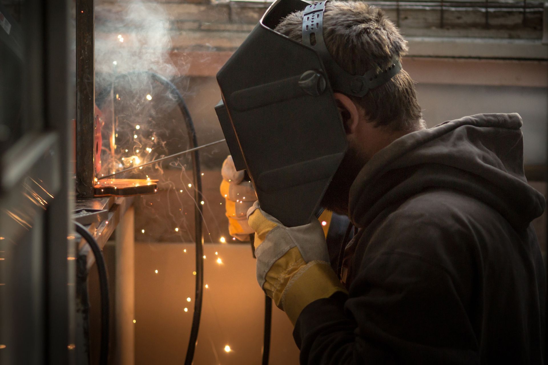 Worker welding metal beside a window, with sparks flying and a glowing orange reflection indoors.