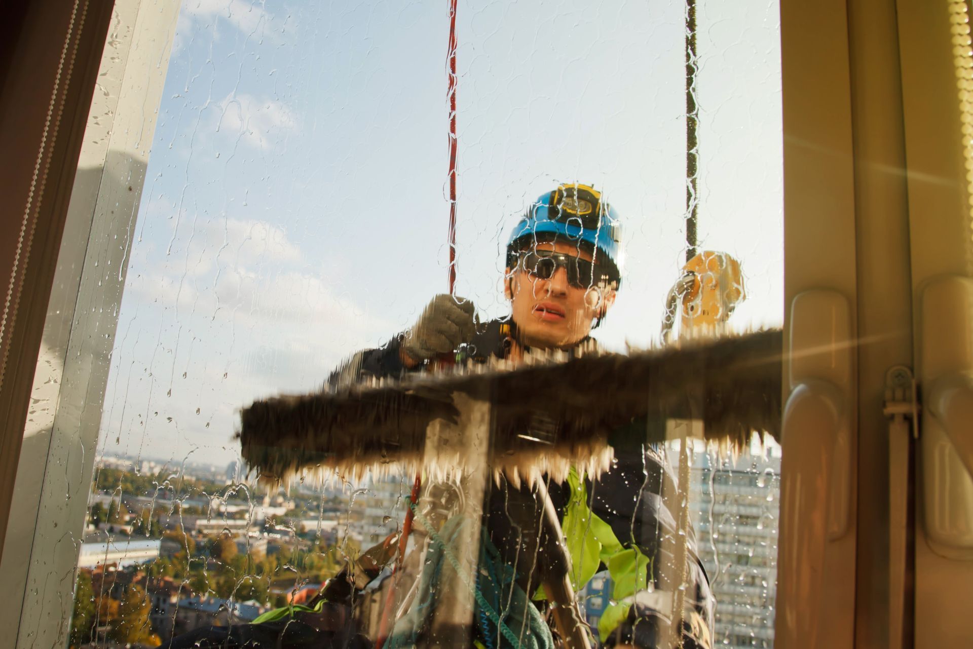 Window cleaner on a high-rise exterior, wiping a glass pane with city skyline behind.