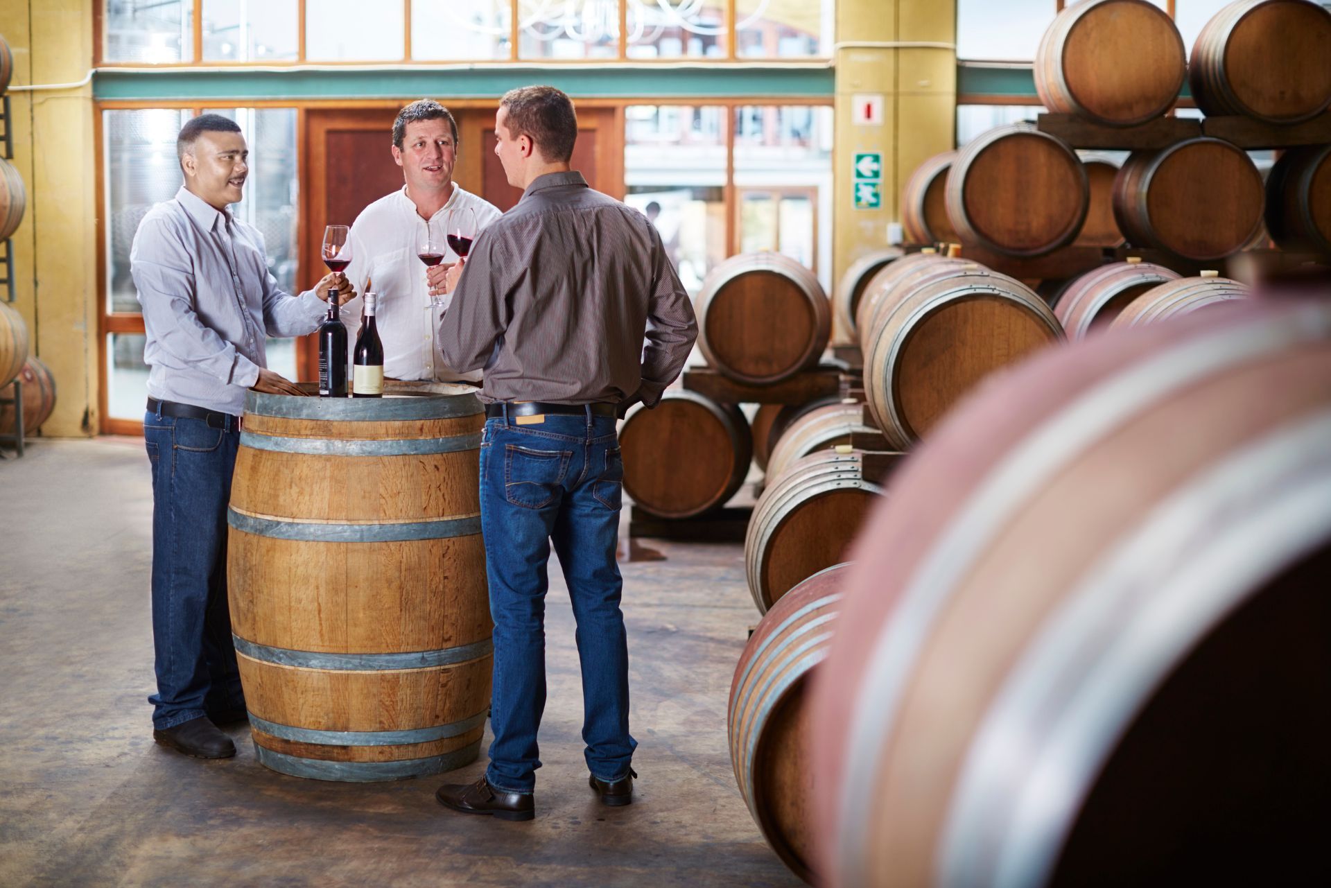 Three people tasting wine beside a wooden barrel in a winery, with stacked barrels in the background.