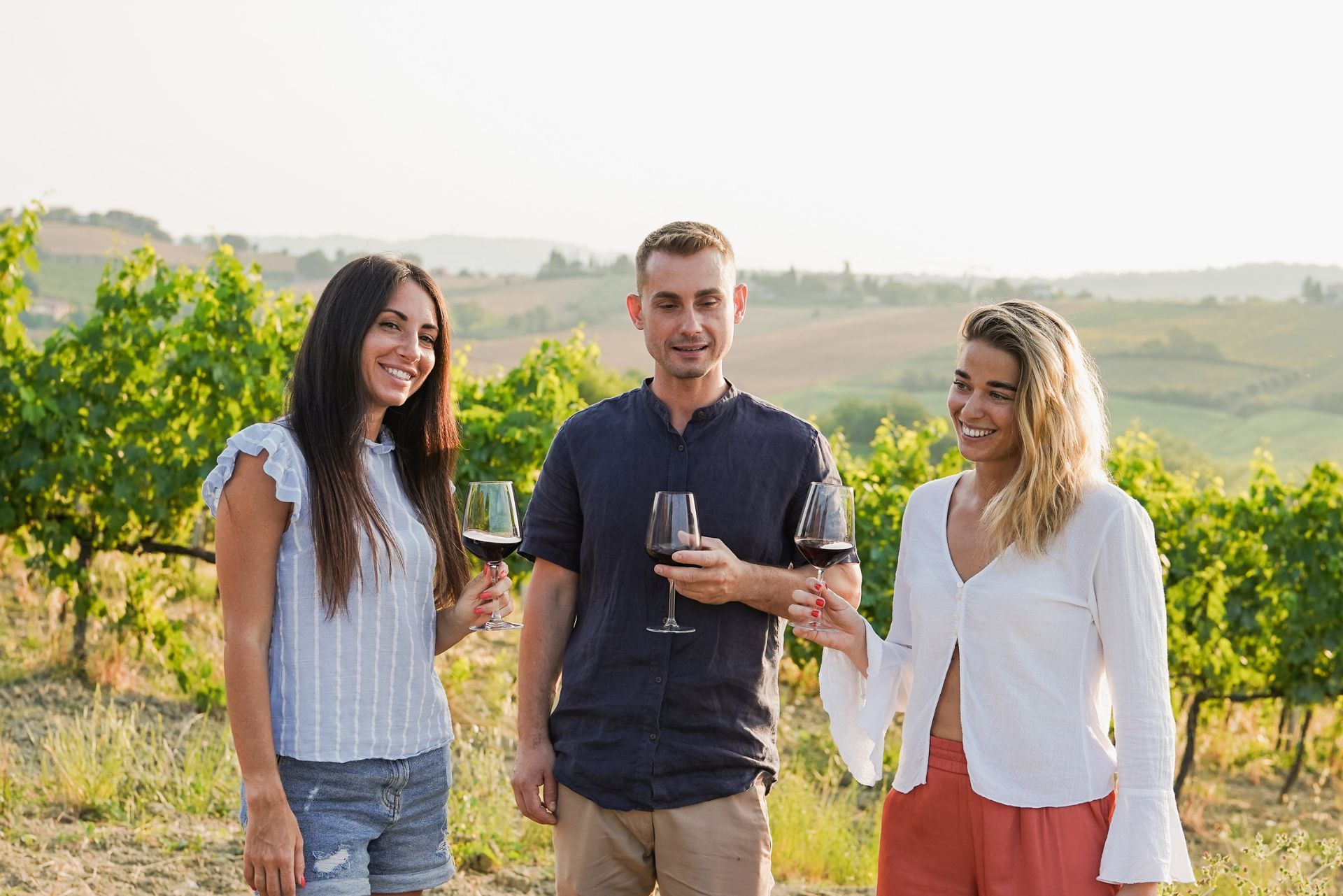Three people smiling with wine glasses in a vineyard at sunset