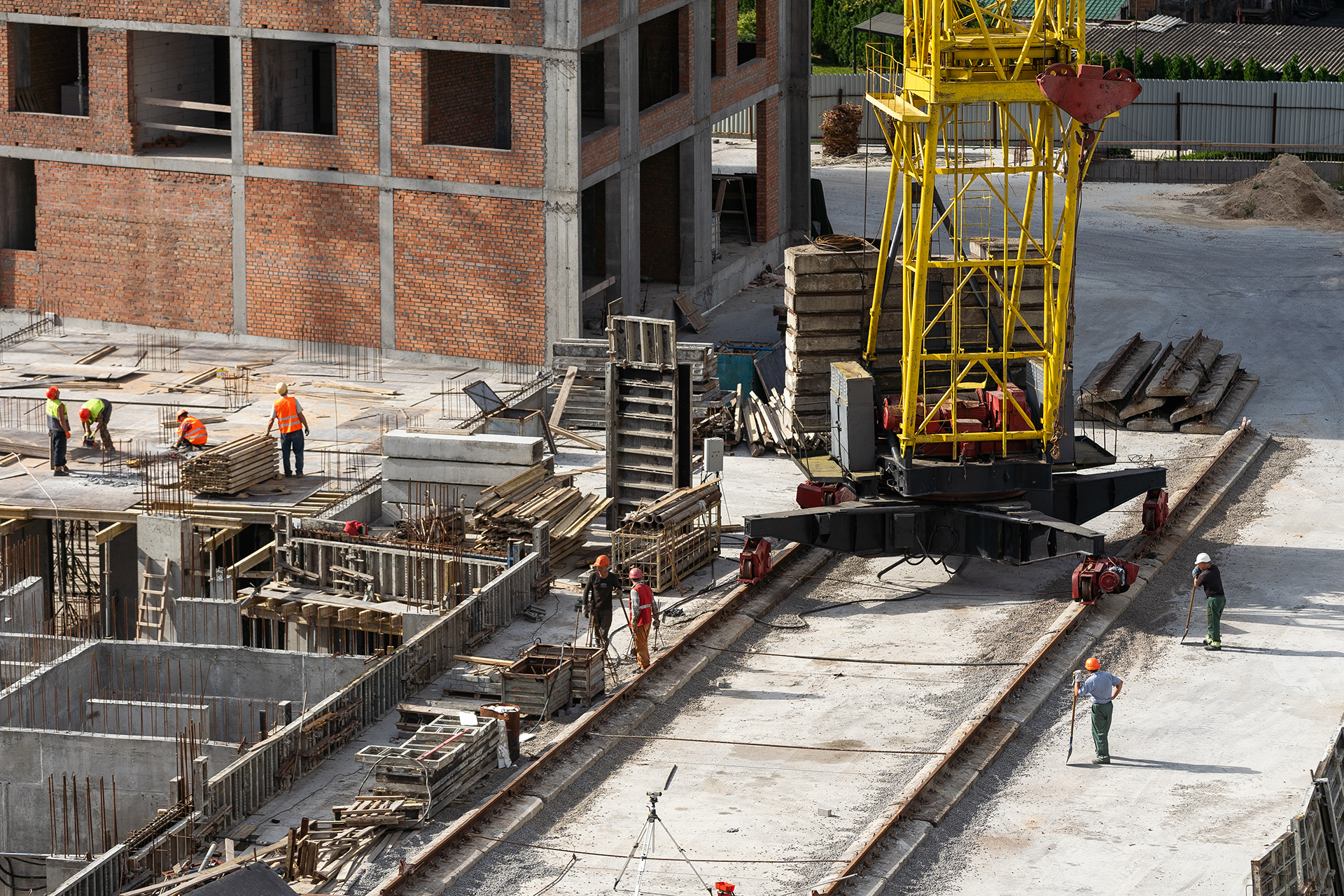 Construction site with workers, concrete foundations, and a yellow crane beside a brick building
