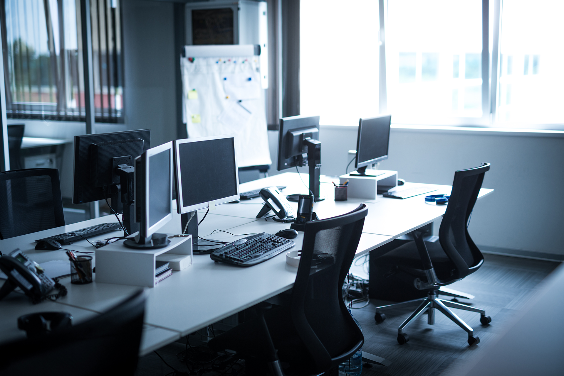 Empty office with desks, computer monitors, and black chairs by a bright window