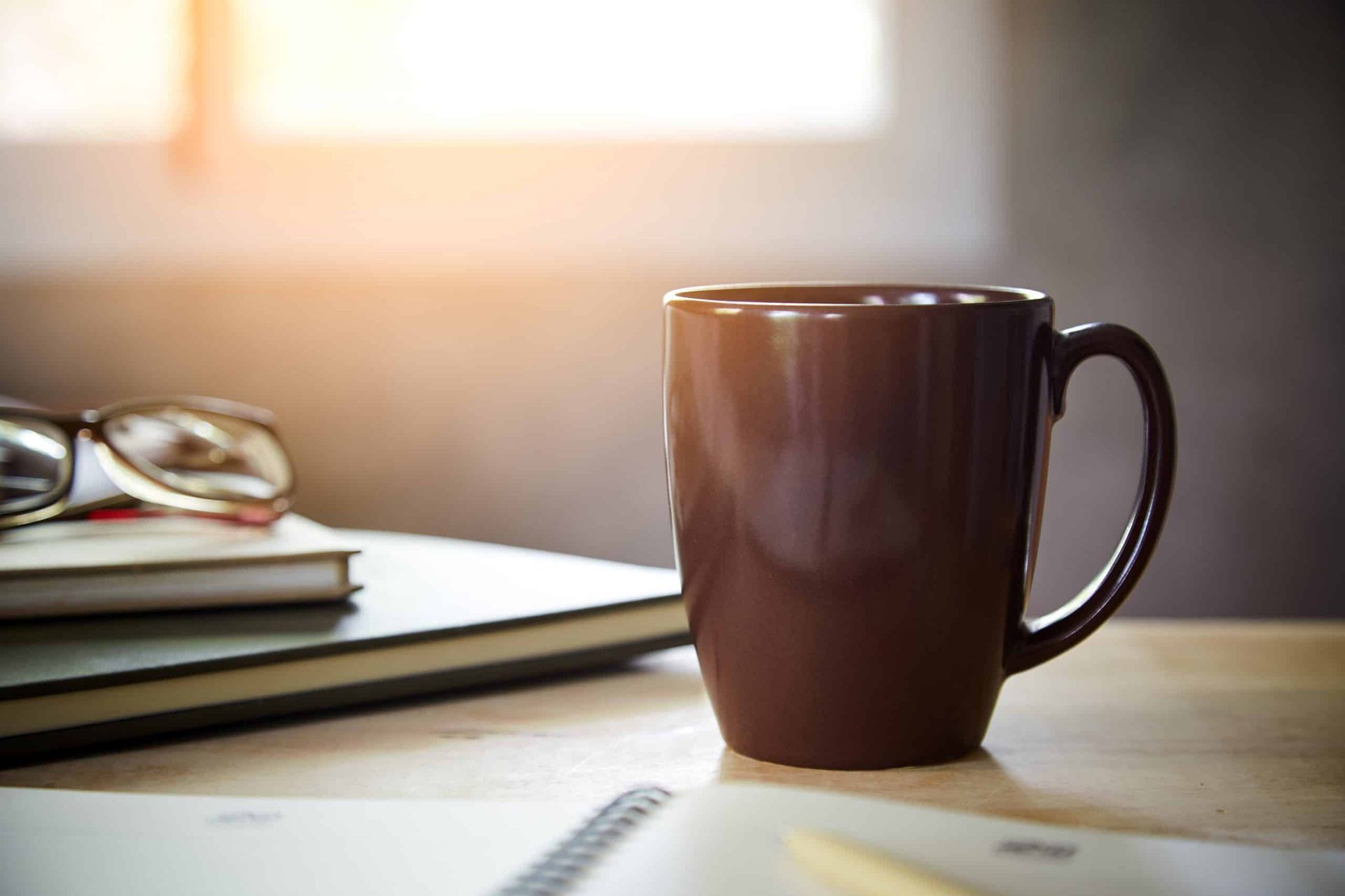 A dark brown mug sits on a wooden desk next to a notebook, pen, and pair of eyeglasses, lit by soft sunlight.
