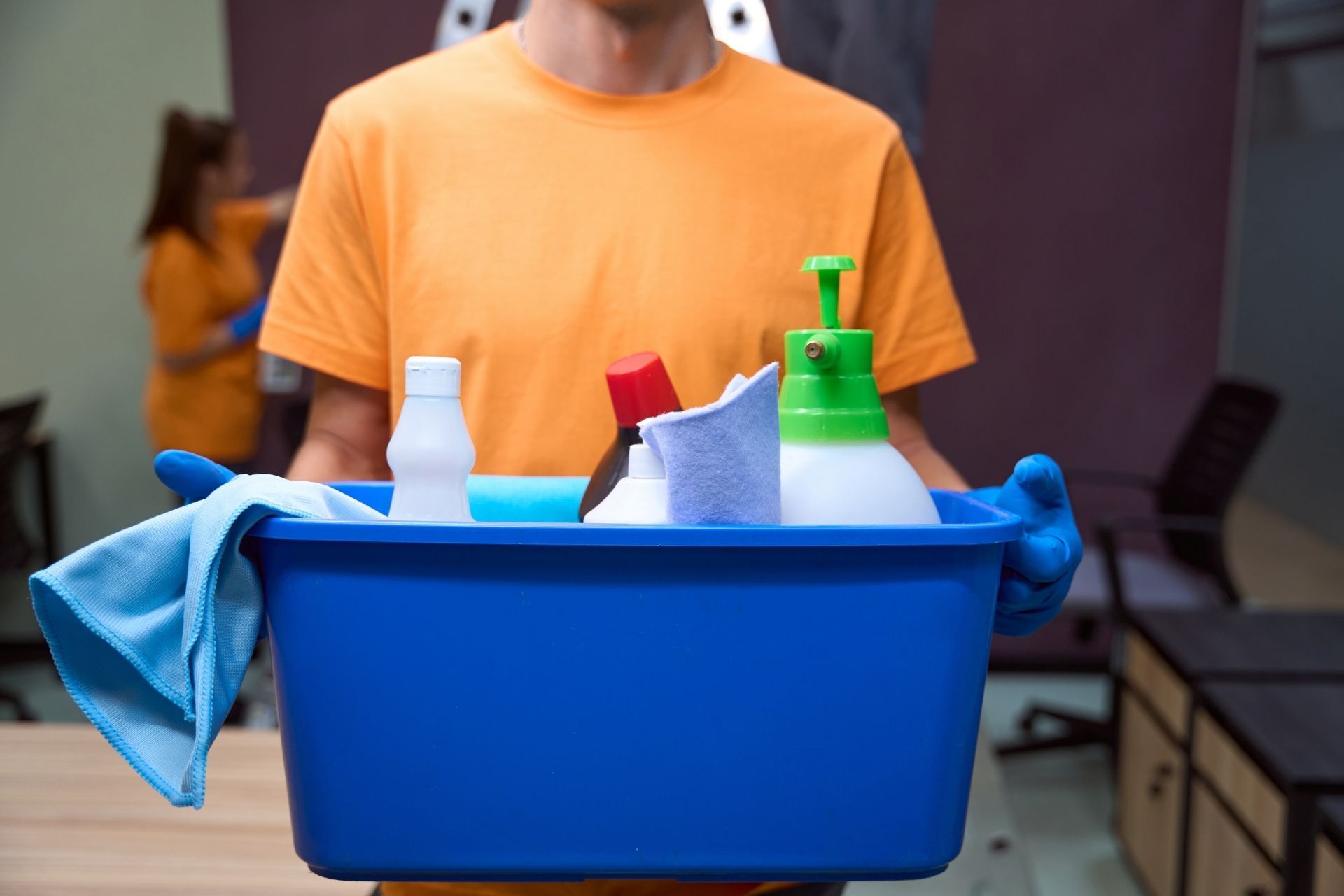 Cleaning supplies in a blue bin held by a person in an orange shirt