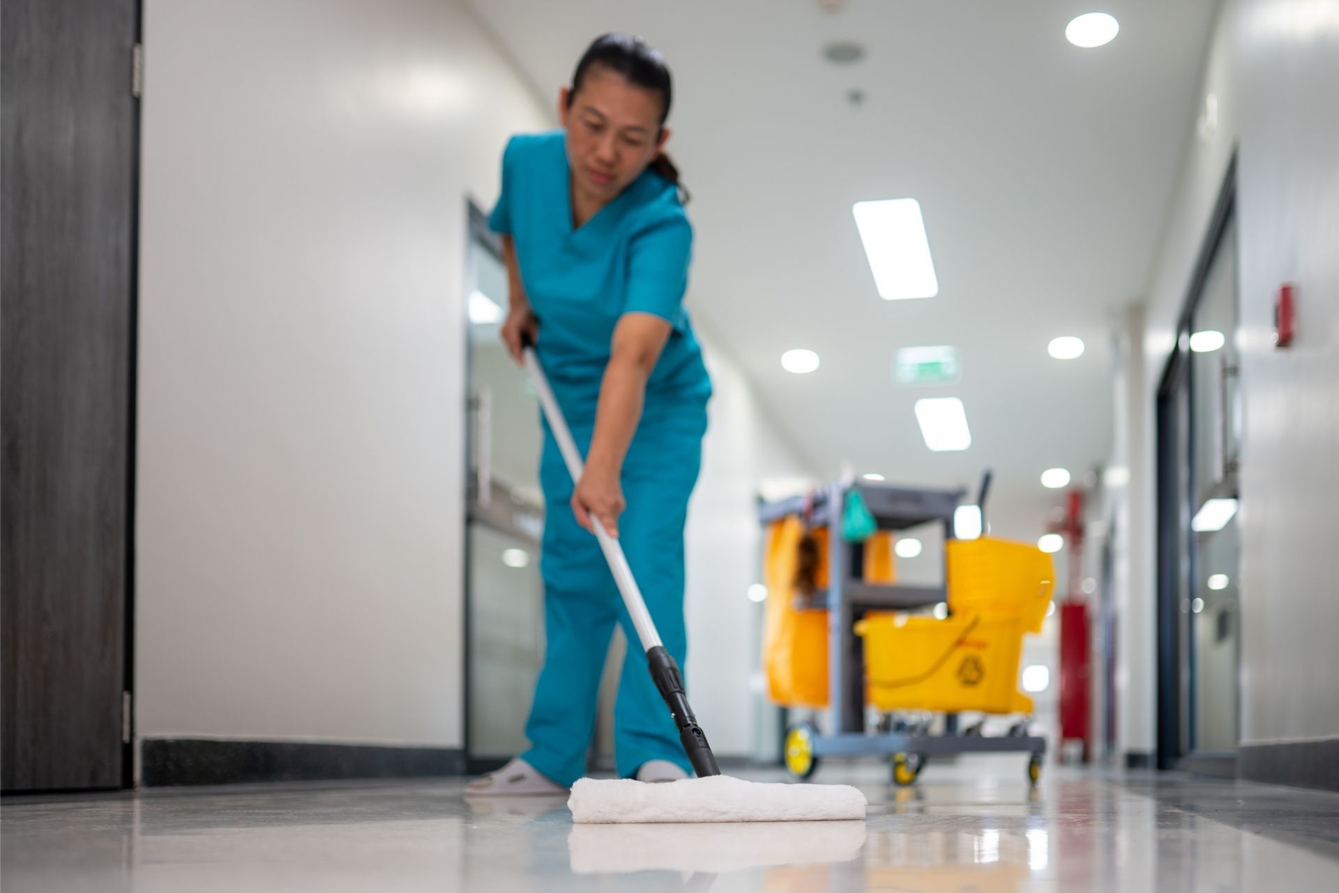 Janitor mopping a hospital hallway floor with a yellow cleaning cart nearby