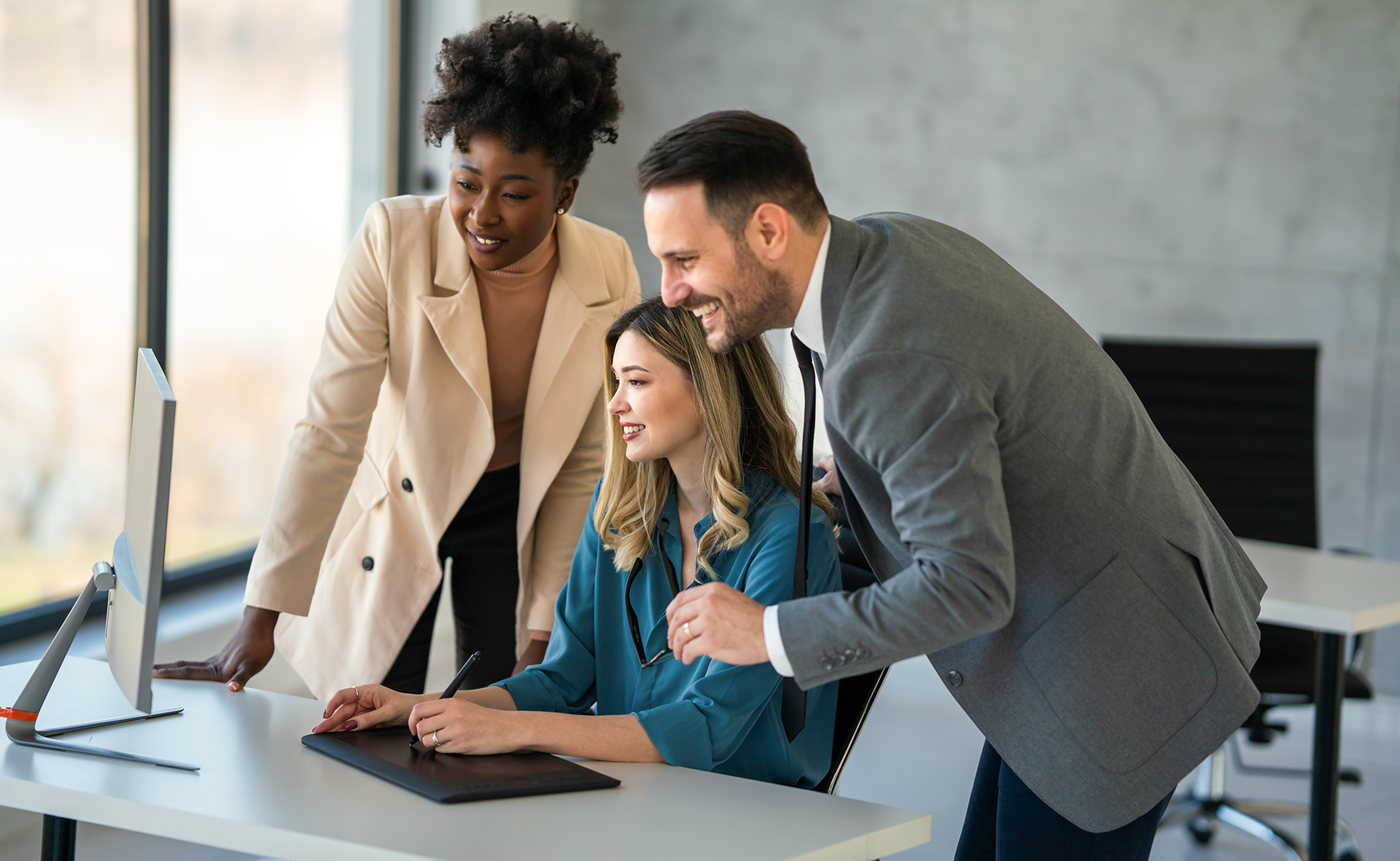 Three coworkers reviewing a laptop together in a bright office, smiling and leaning in.