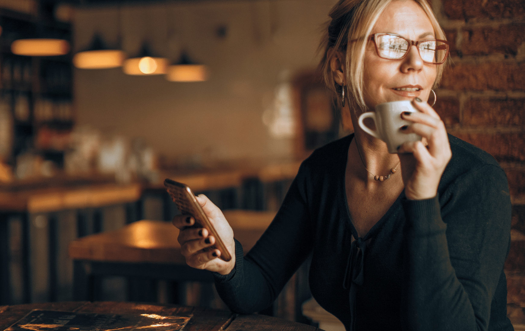 Woman in glasses holding a phone and coffee cup in a warm café, looking off to the side