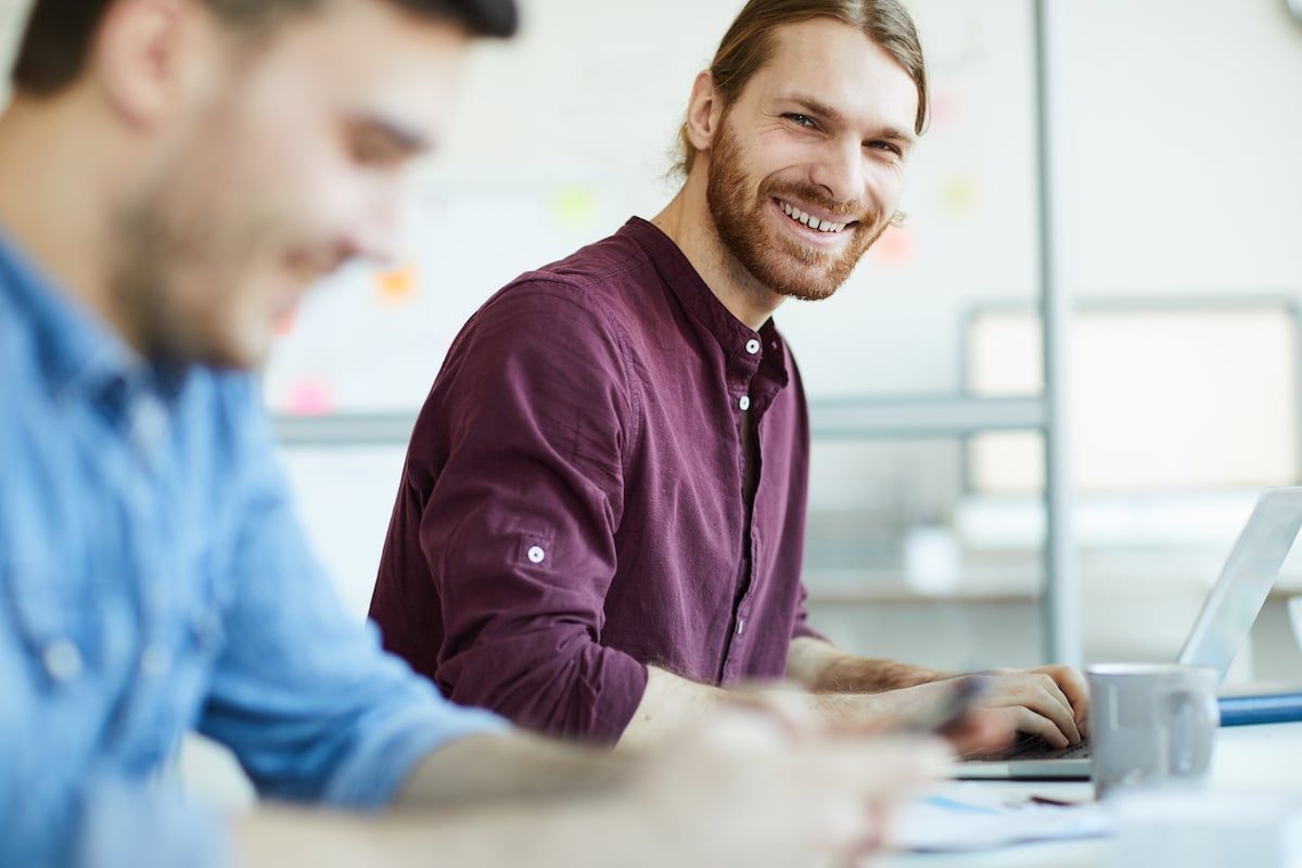 Two coworkers at a bright office table, one smiling while using a laptop and holding a mug