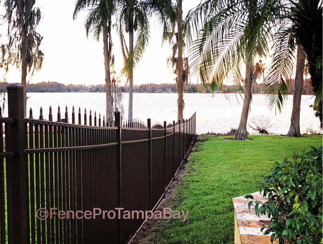Brown metal fence along a green lawn, overlooking a lake with trees and sunset.