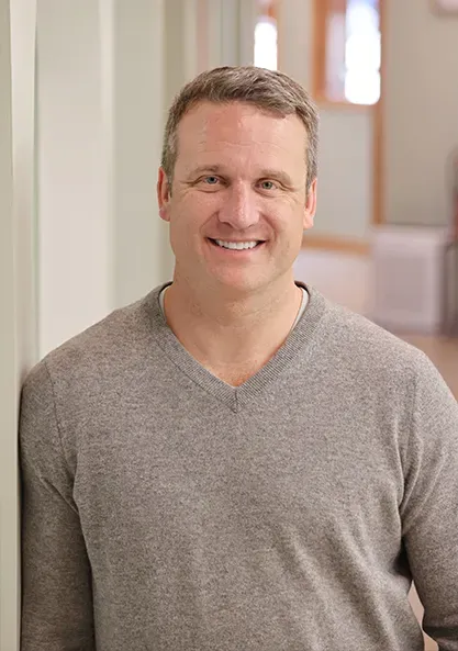 Smiling man in a gray sweater standing indoors in a softly lit hallway