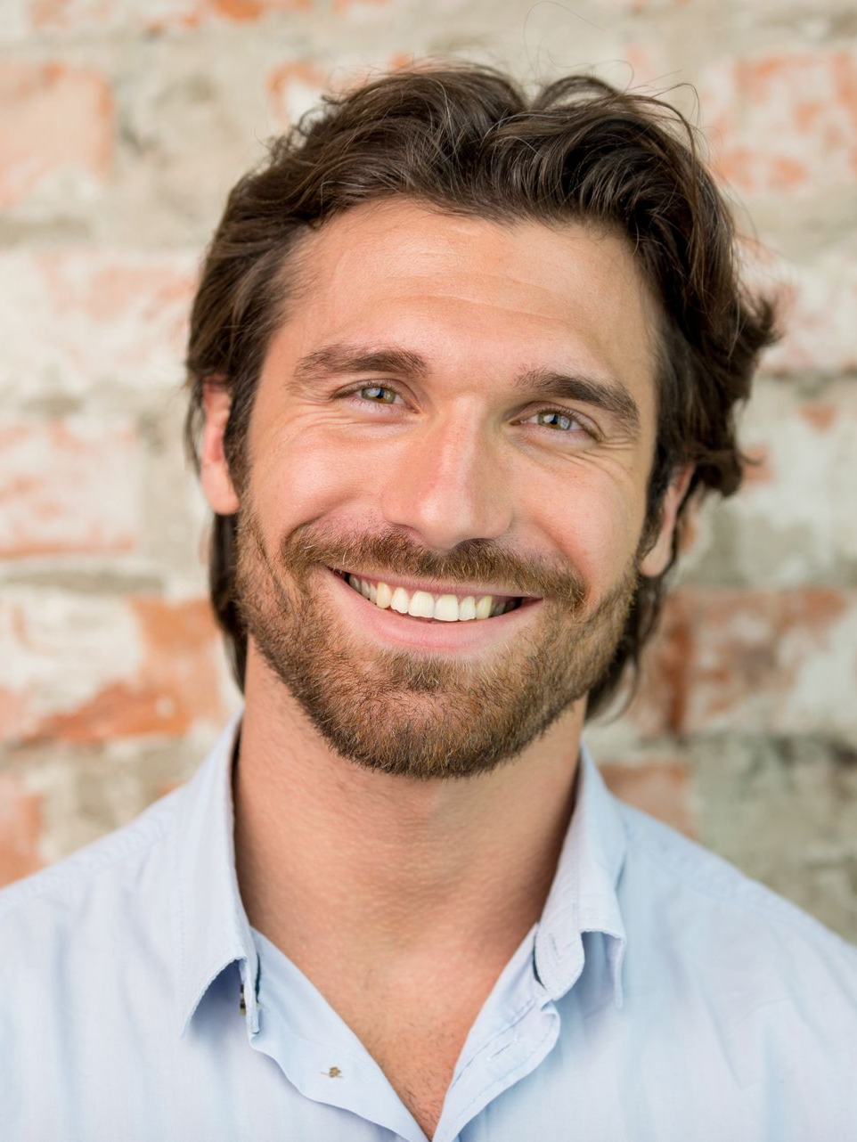 Smiling man with dark hair and beard, wearing a light blue shirt, against a brick wall background