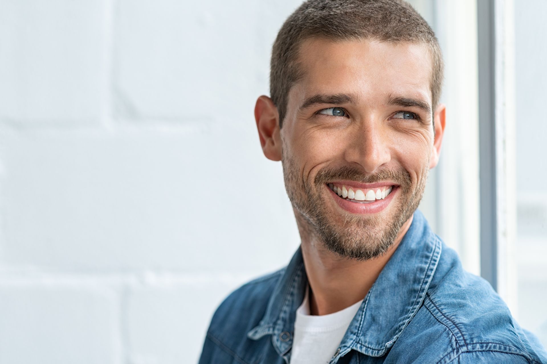 Smiling man in a denim shirt standing by a bright window