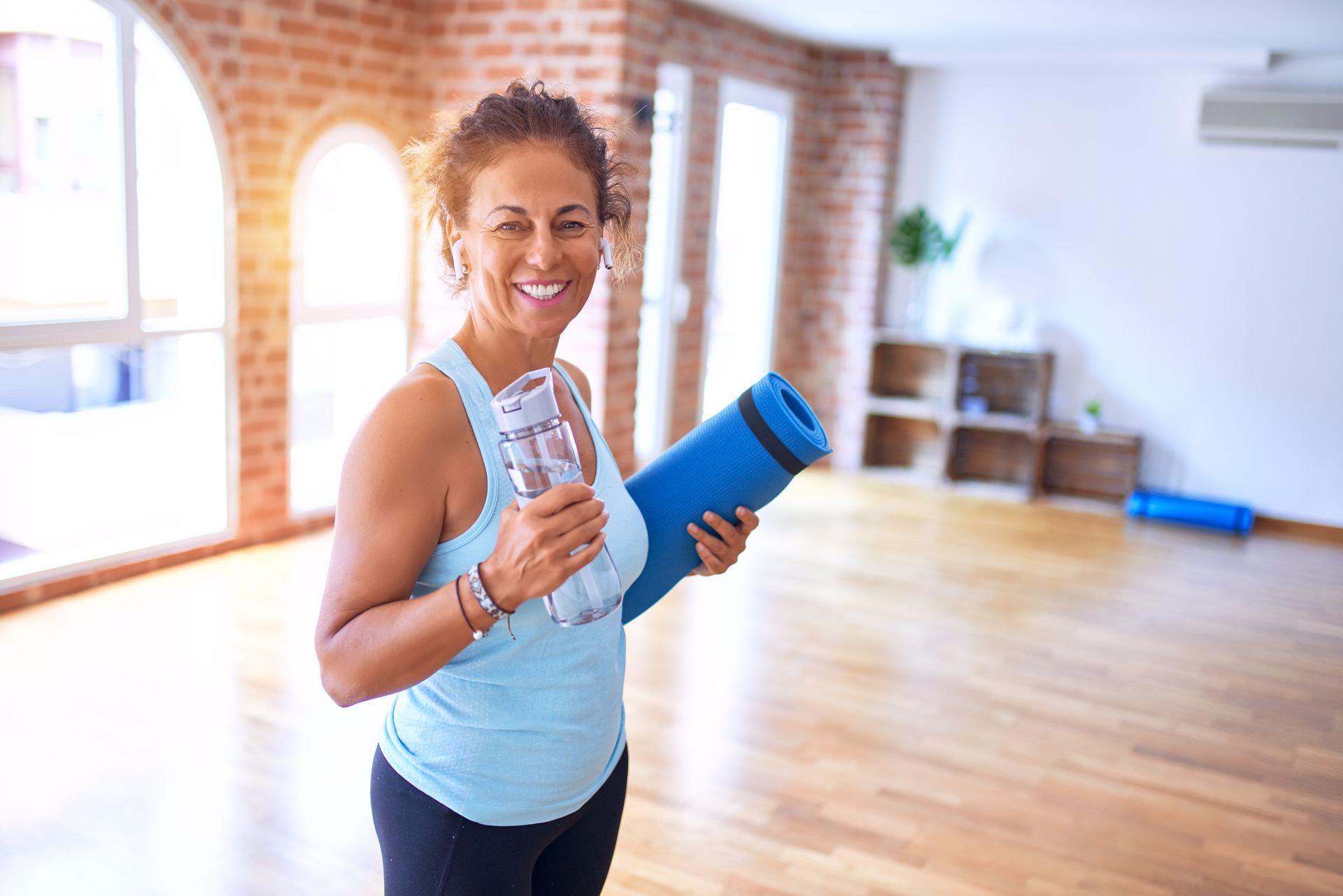 Smiling woman in a bright studio holding a water bottle and blue yoga mat after a workout