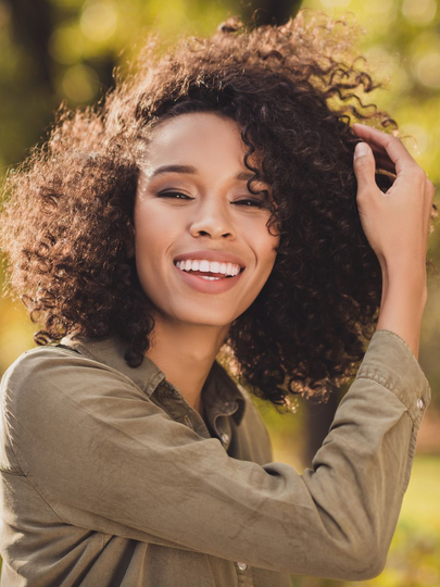 Smiling person with curly hair outdoors, hand touching hair in warm sunlight
