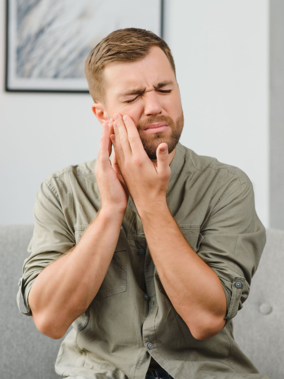 Man sitting on a couch, pressing his cheek with a pained expression.