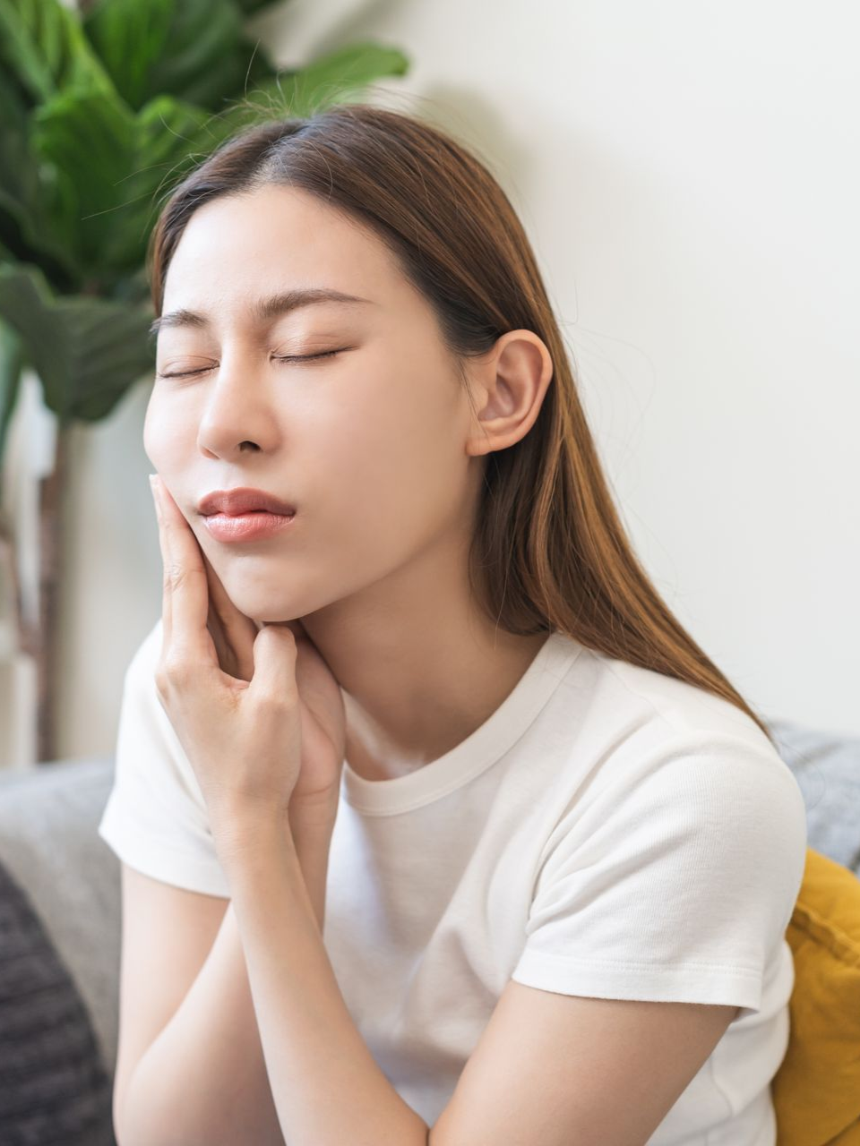 Woman in a white shirt gently touching her cheek, eyes closed, with a pained expression.