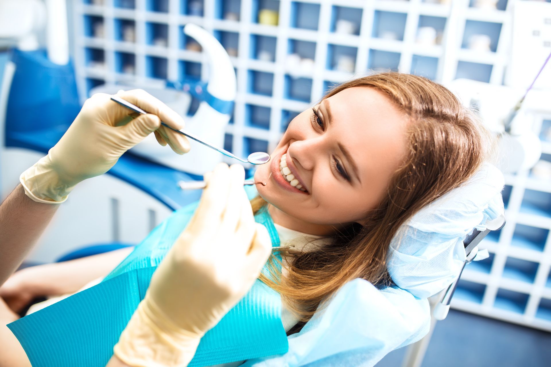 Smiling dental patient in a blue chair while a dentist holds tools in a bright clinic