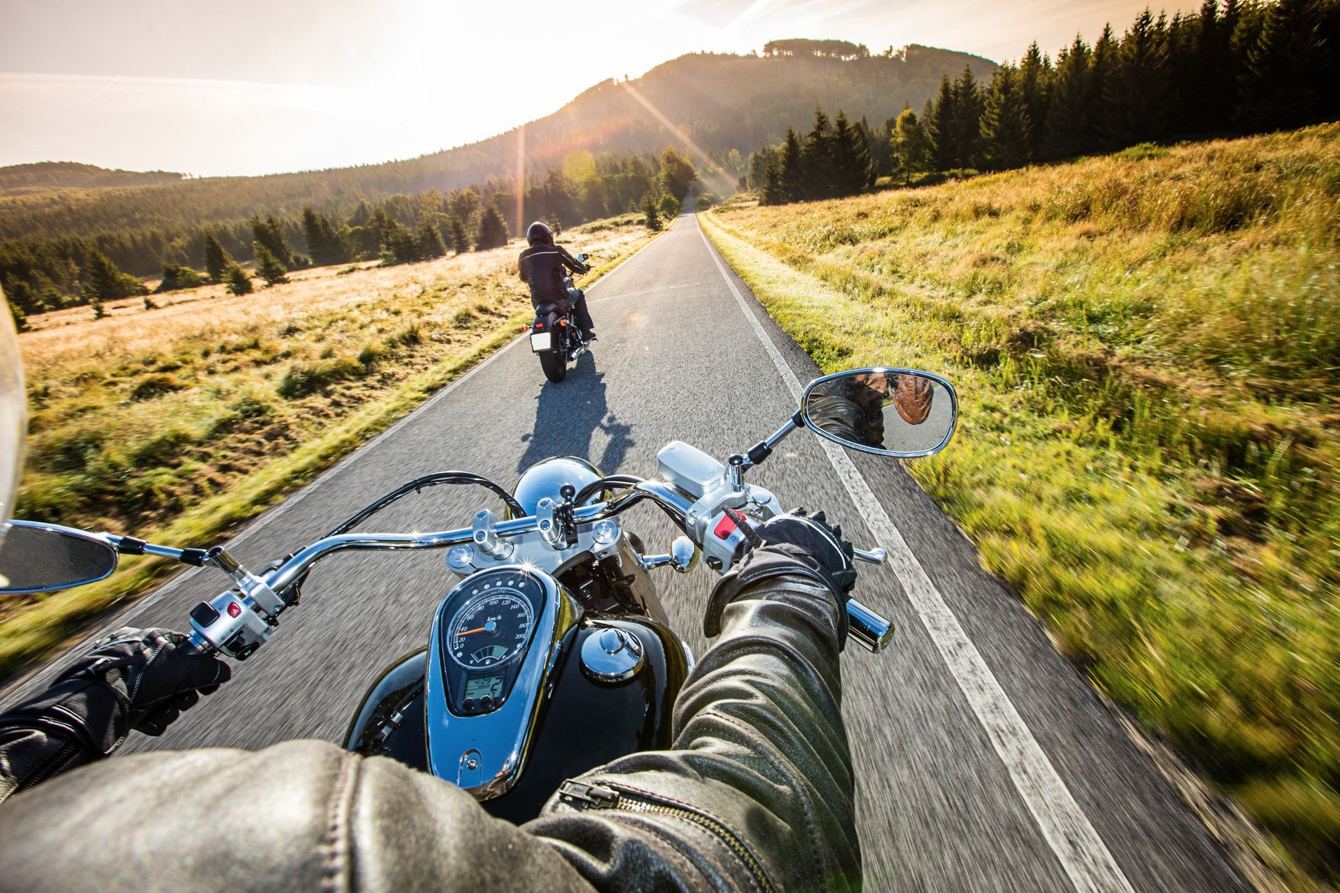 Motorcyclists riding a mountain road at sunset, viewed from behind a blue bike with wide handlebars