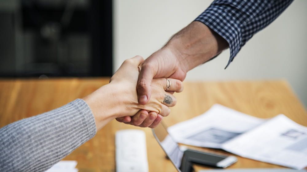 Dos personas dándose la mano sobre un escritorio con papeles y un ordenador portátil al fondo.